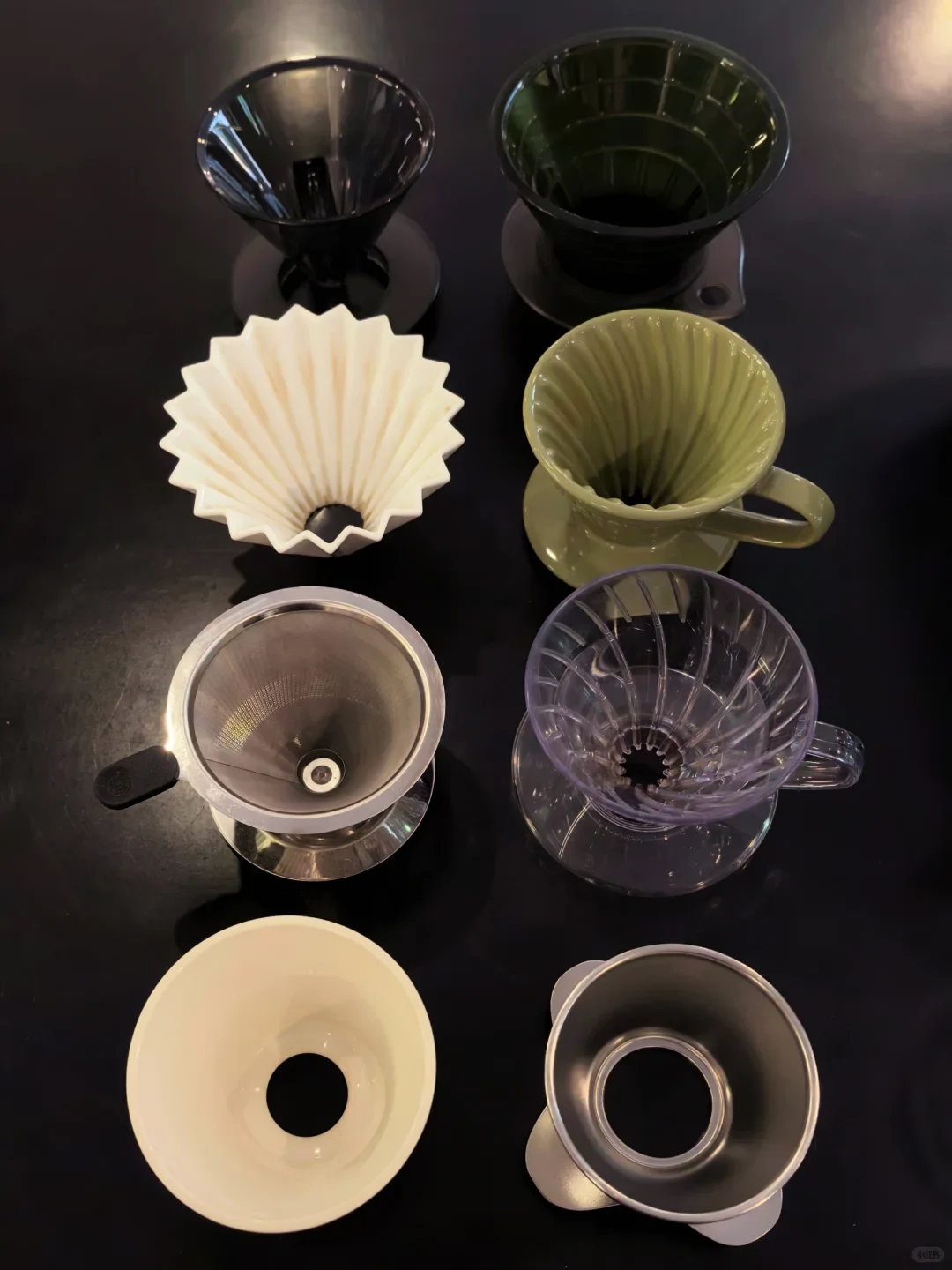 A top-down photo shows eight pour-over coffee drippers and filters in assorted materials and colors arranged in two columns on a dark tabletop.