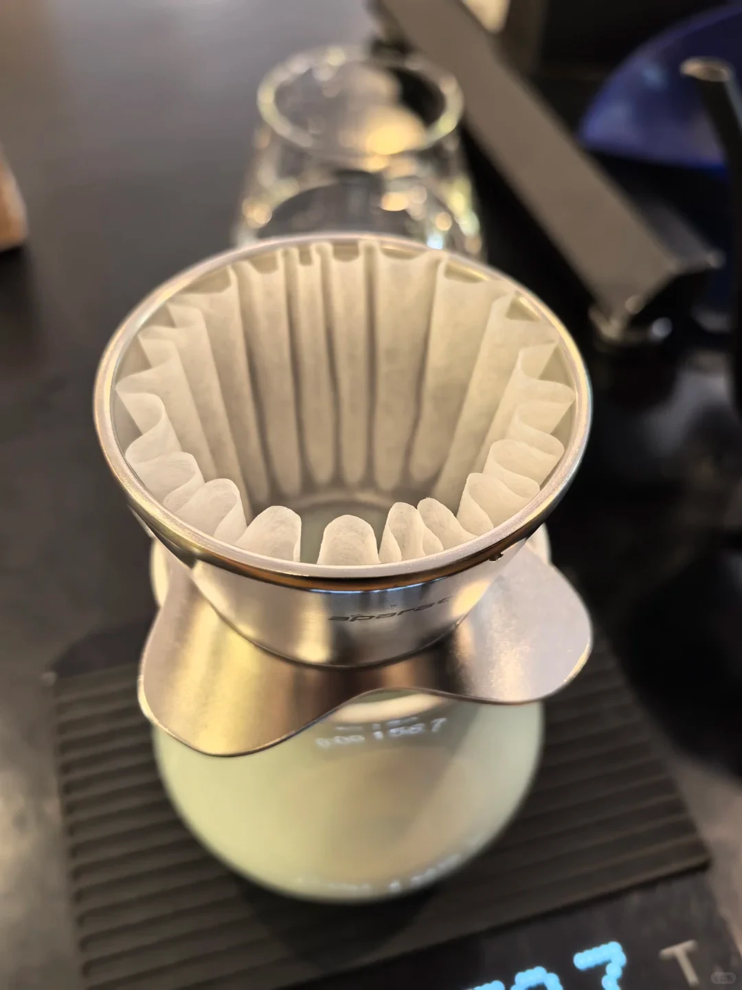 A silver metal dripper lined with wave paper sits over a rounded pale-green vessel, with a scale display below. Its exact model is not established by the photograph.