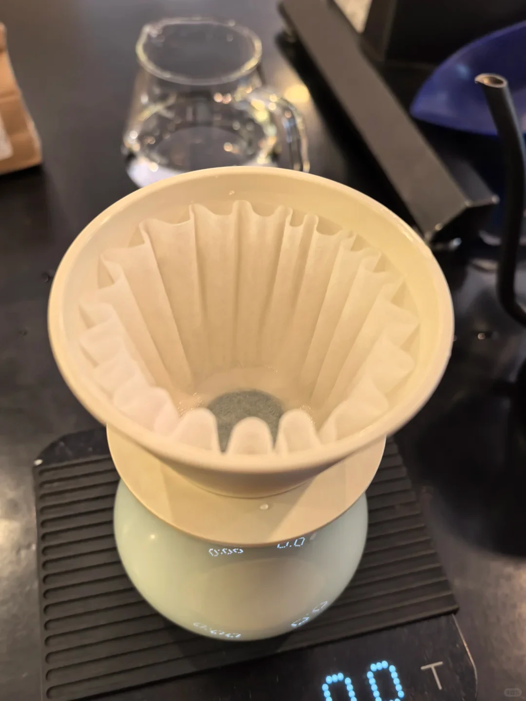 A cream-colored dripper holds a wave paper filter above a pale-green rounded vessel, with illuminated scale readings visible below.