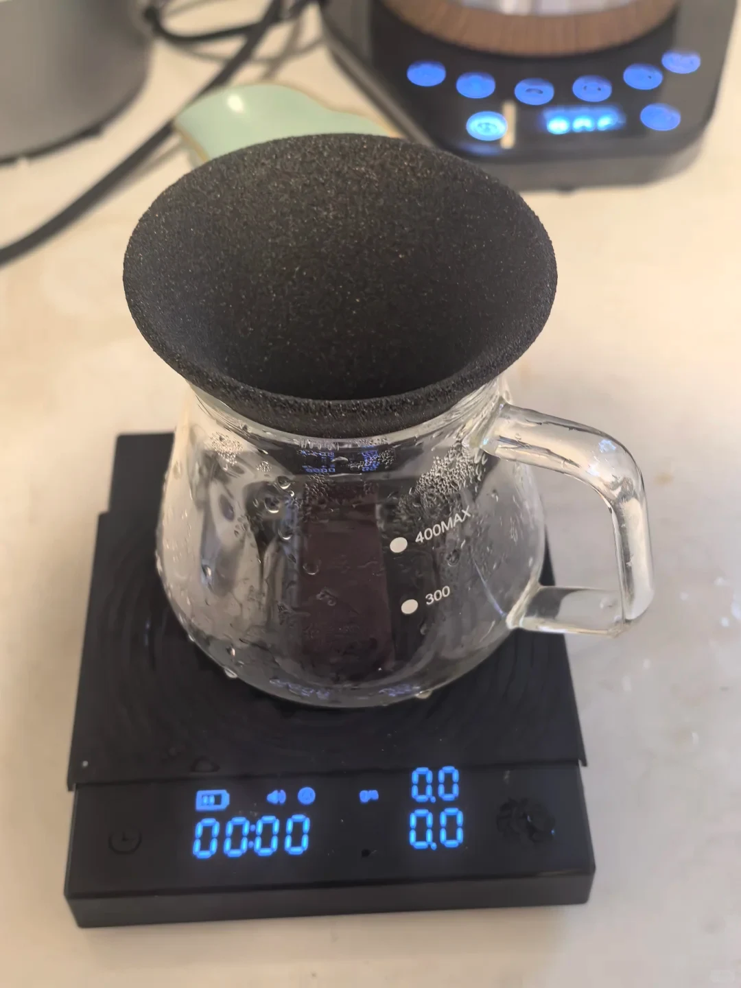 A clear glass coffee server with a black dripper on top sits on a black digital scale; the server is marked “400MAX” and “300,” and the scale display reads 0.0.
