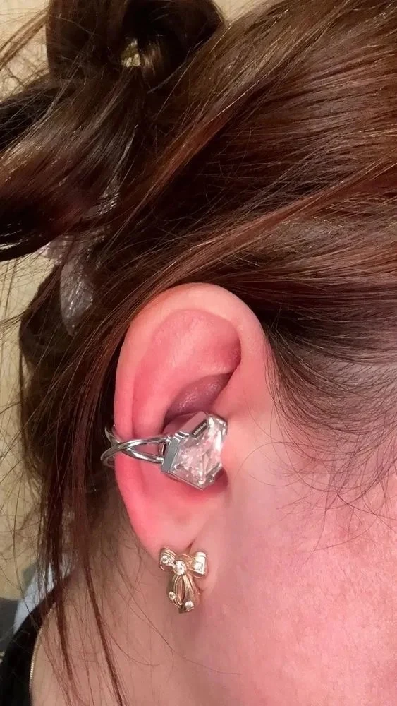 Close-up of a person's ear wearing a clip-on wireless earbud styled with an oversized faceted faux-diamond design, along with a small bow-shaped earring on the earlobe.