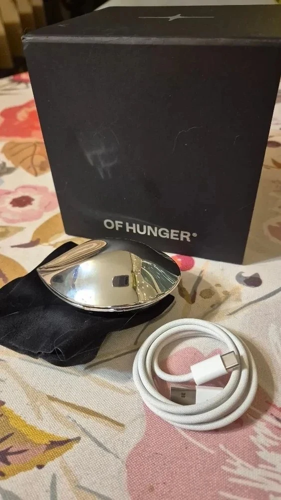 A metallic chrome pebble-shaped device resting on a black velvet pouch in front of a black box branded OF HUNGER, alongside a coiled white braided USB cable on a patterned fabric surface.