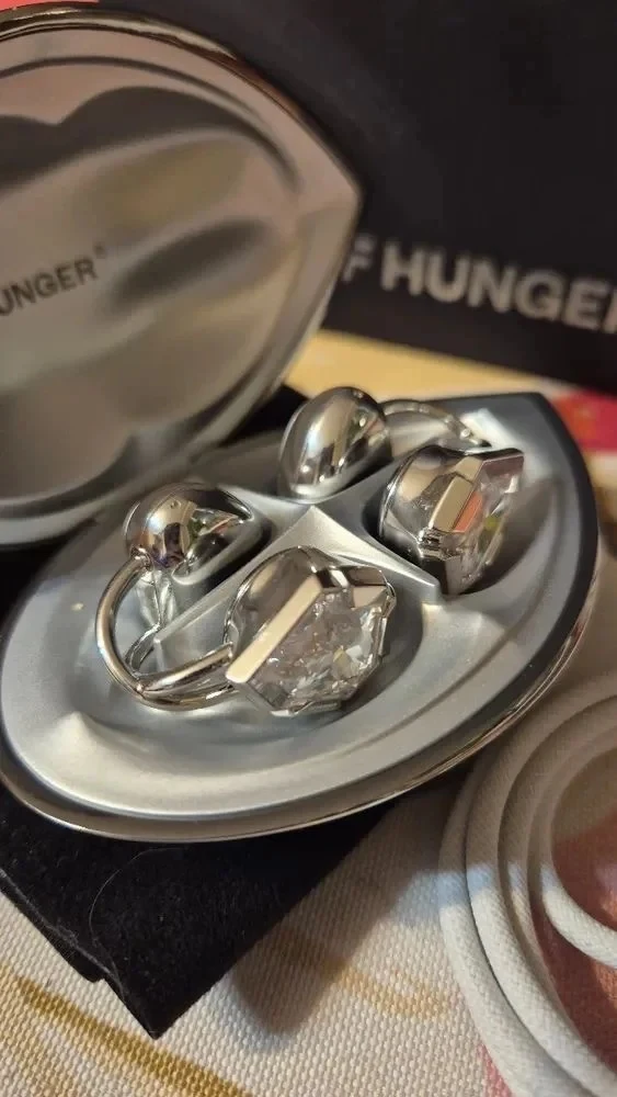 An open silver-tone charging case displaying a pair of clip-on wireless earbuds designed with faceted clear crystal accents and metallic finishes.