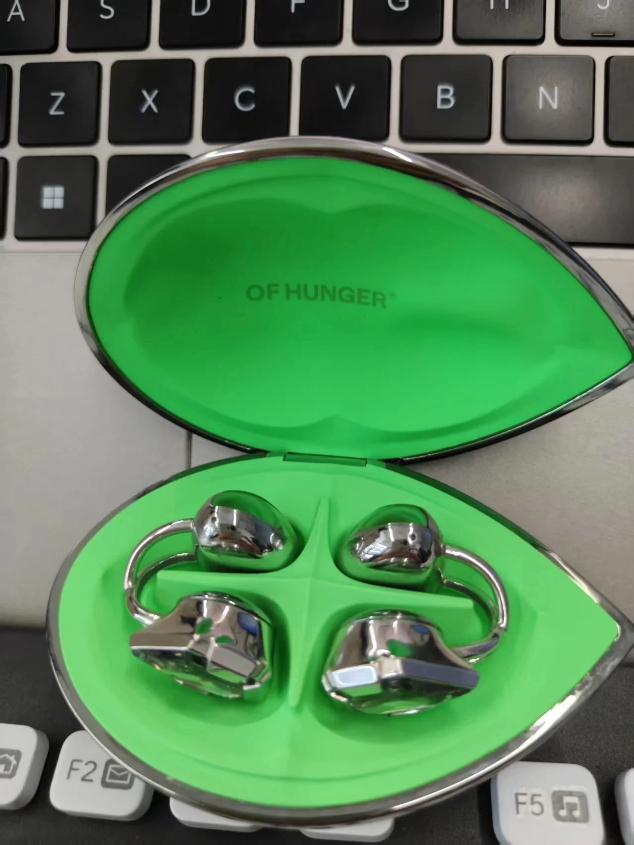 An open metallic teardrop-shaped charging case with a bright green interior resting on a laptop keyboard, holding a pair of chrome-finished clip-on wireless earbuds, with "OF HUNGER" printed inside the lid.