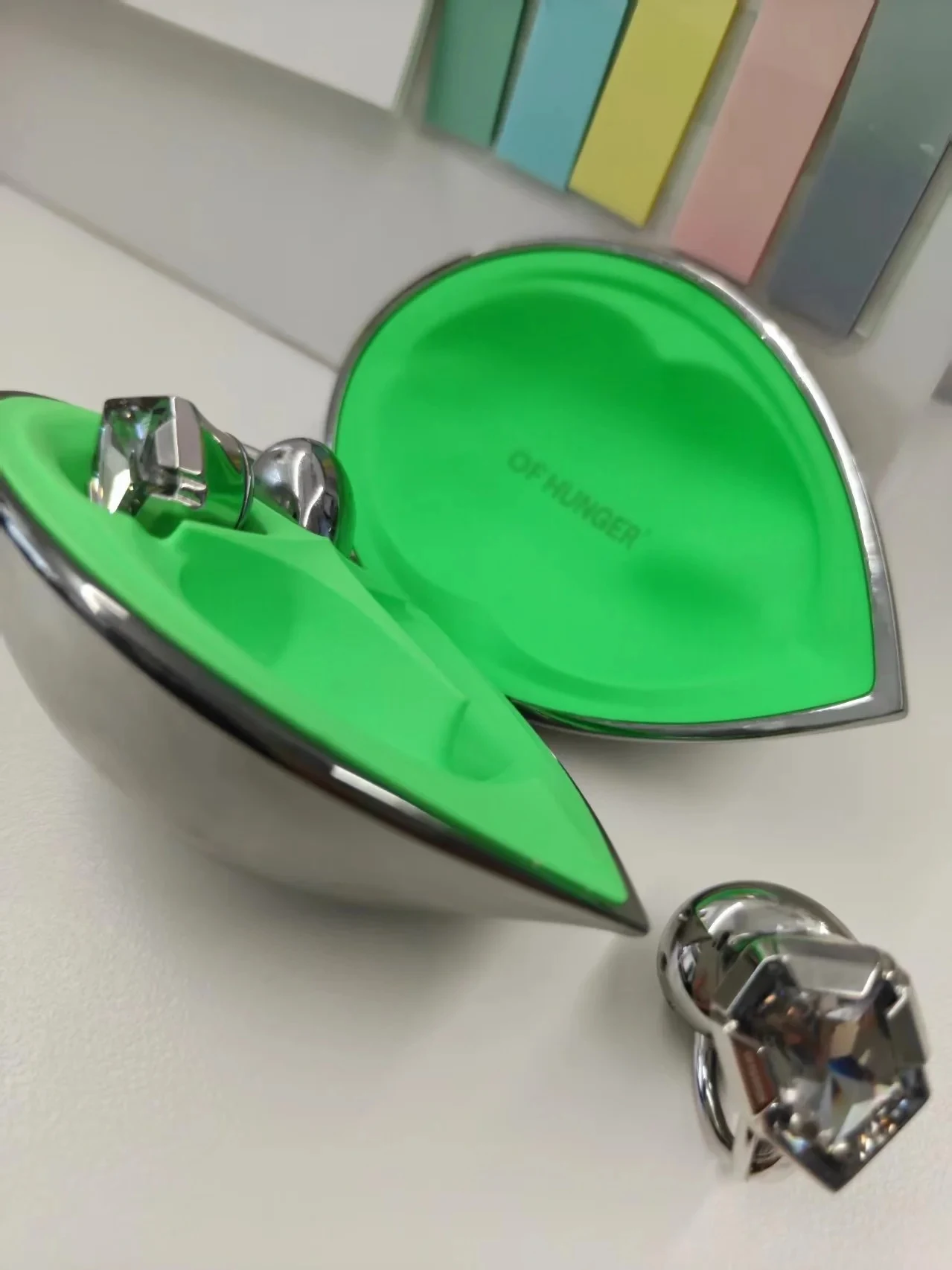 An open metallic teardrop-shaped charging case with a vivid neon-green interior labeled "OF HUNGER," containing one metallic ring-style earbud with a faceted crystal, while the matching earbud sits nearby on a white surface.