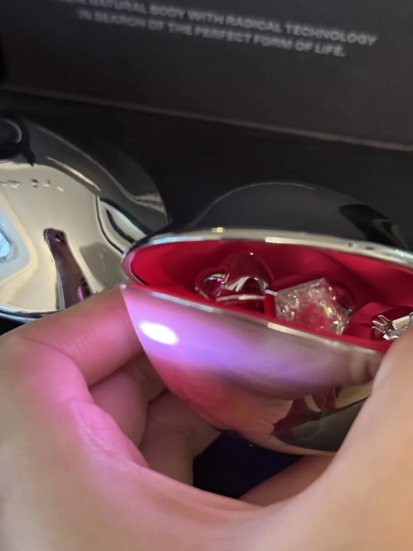 A hand holds open a shiny metallic capsule case lined with hot pink fabric, revealing silver jewelry items inside.