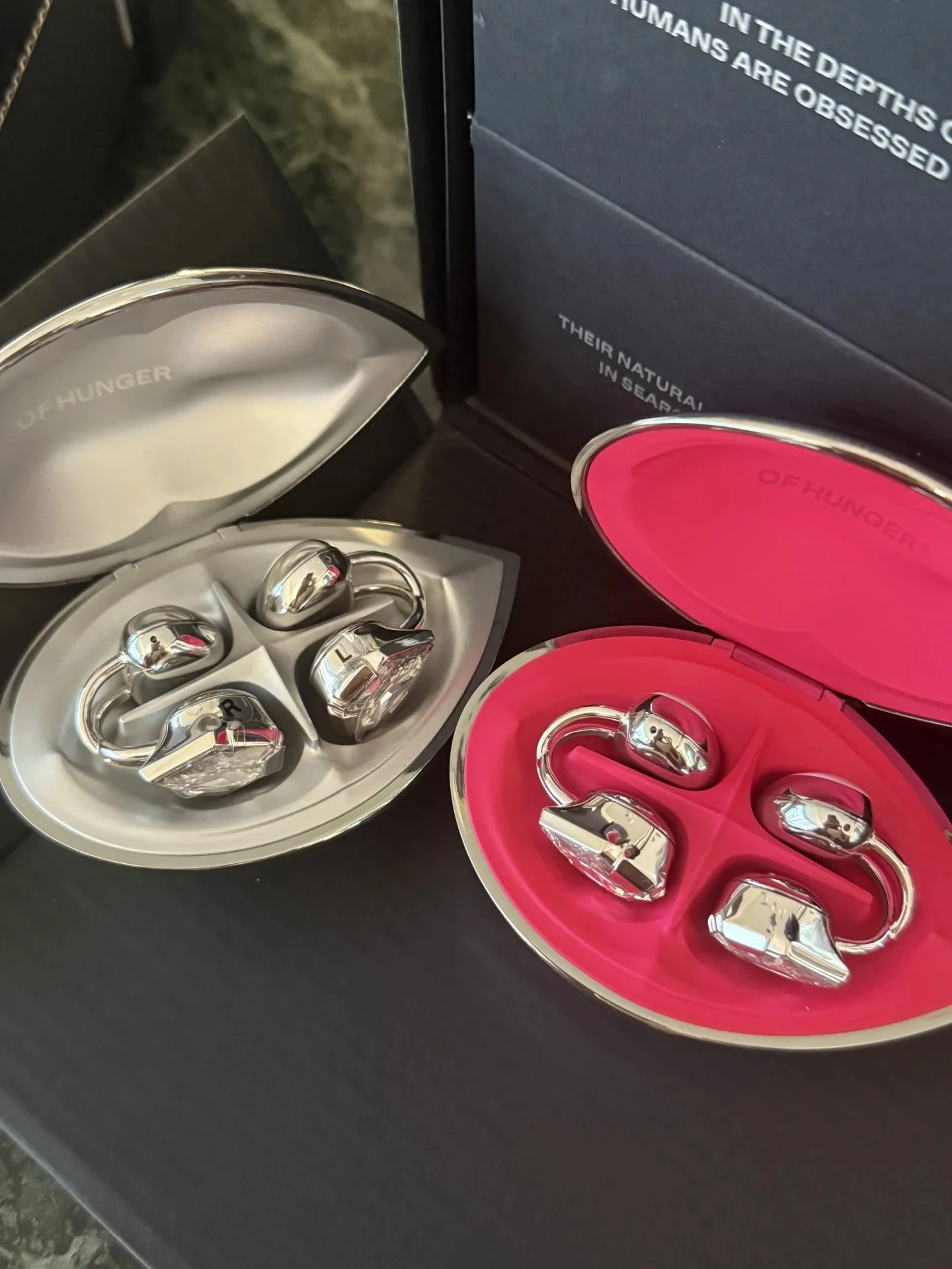 Two pairs of silver ear-cuff style earbuds resting in open, lip-shaped charging cases—one with a silver interior and the other with a vibrant pink interior—set against dark packaging.