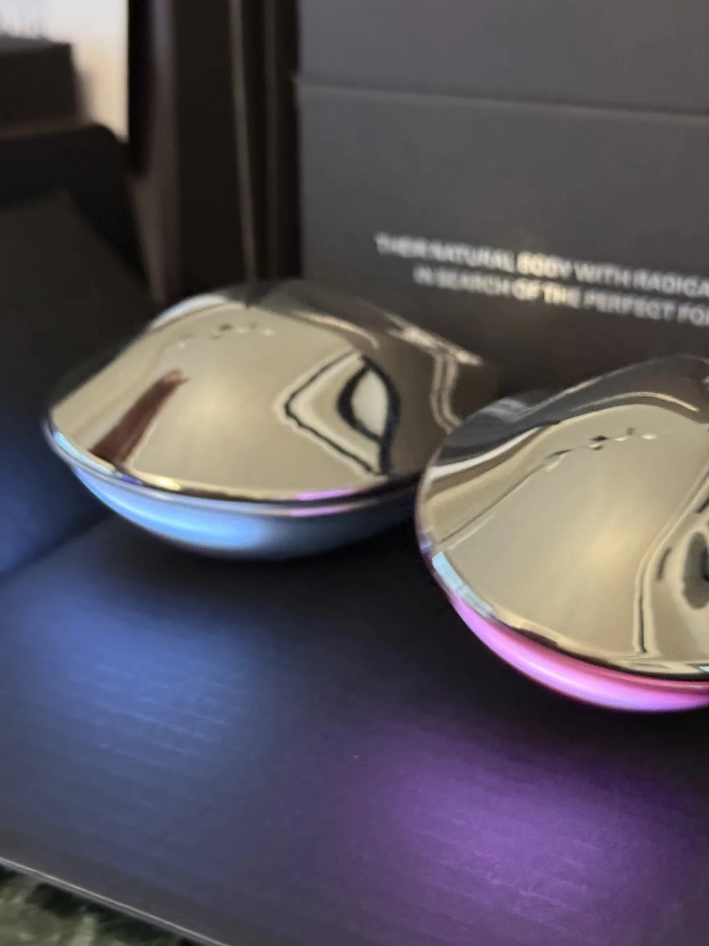 Two metallic chrome scalp massage brushes resting inside dark packaging, one featuring a silver-blue base and the other a pink base.