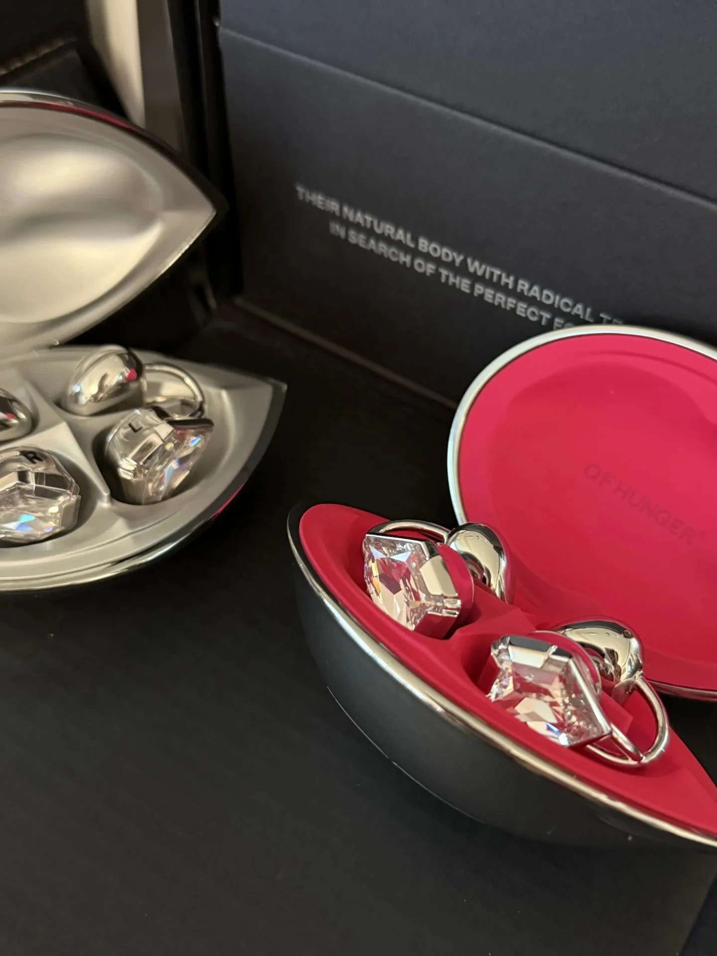 Two metallic cases holding decorative crystal-studded ear accessories or open-ear earphones, one case lined in bright pink embossed with 'OF HUNGER' and another lined in silver.