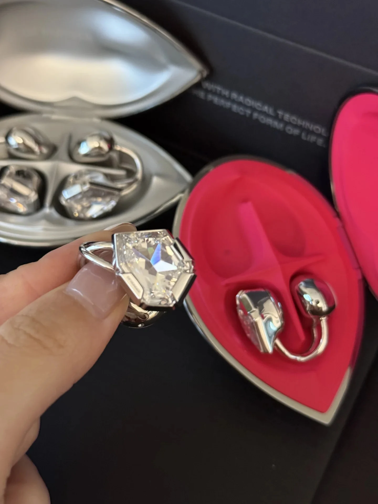 A hand holds a futuristic silver ear accessory featuring a large geometric faceted clear crystal, set in front of open metallic and bright pink teardrop-shaped cases holding matching jewelry.
