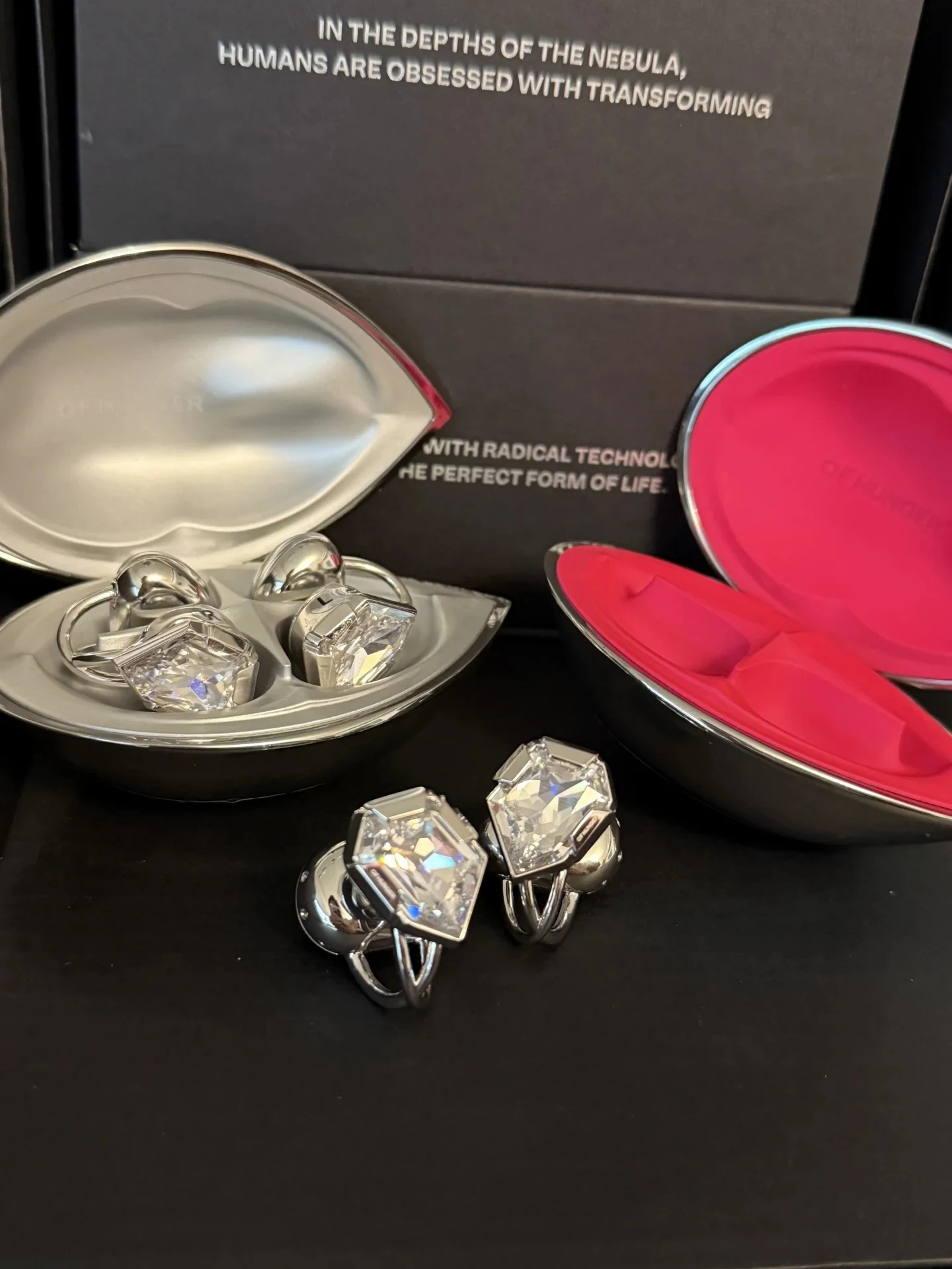 Silver ear clips with large, faceted clear crystals displayed beside and inside two lip-shaped metallic cases, one with a silver interior and one with a hot pink interior, set against black packaging with English text.