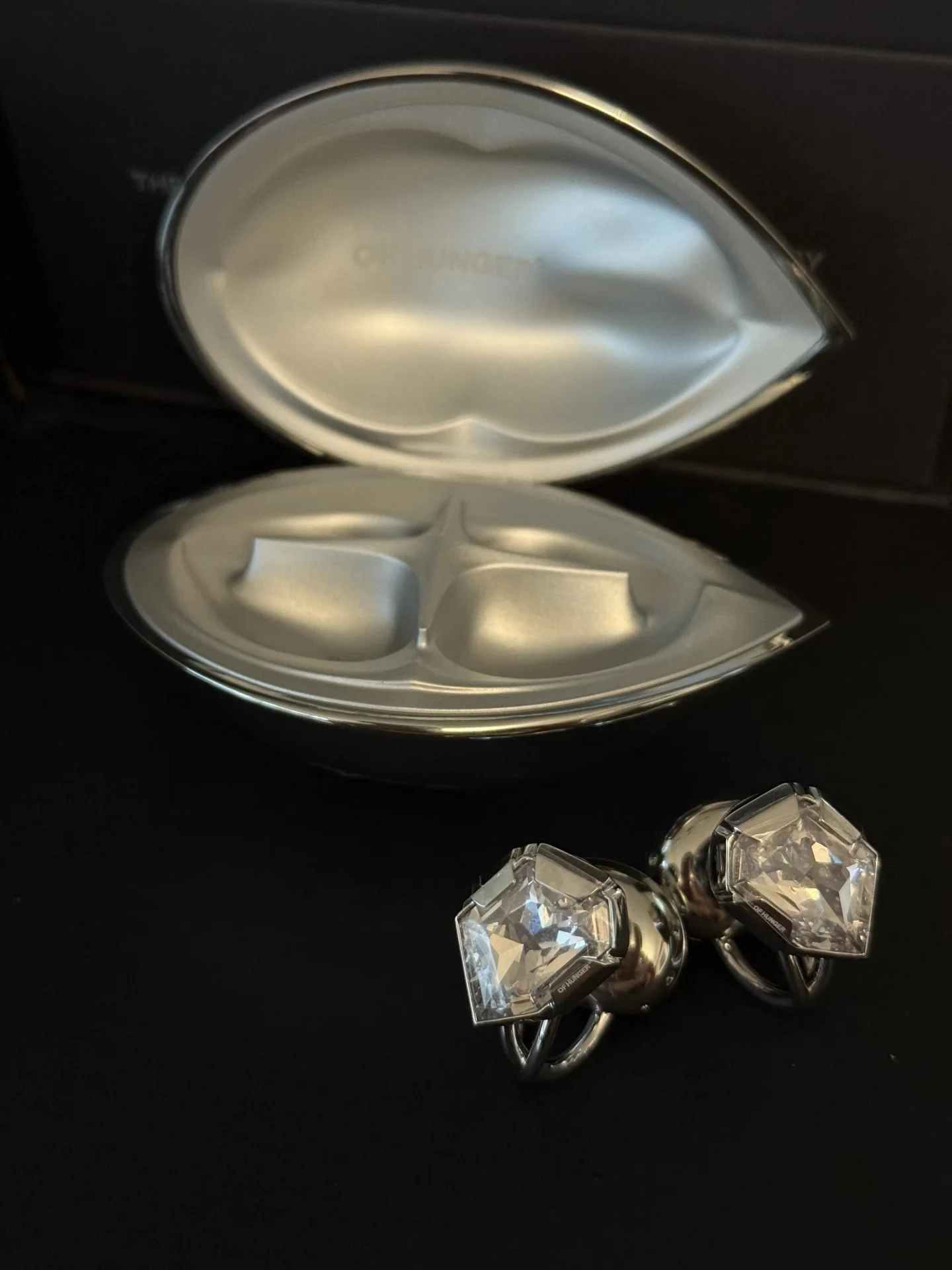 A pair of silver jewelry pieces with large faceted clear crystals beside an open silver metallic case shaped like stylized lips on a black surface.