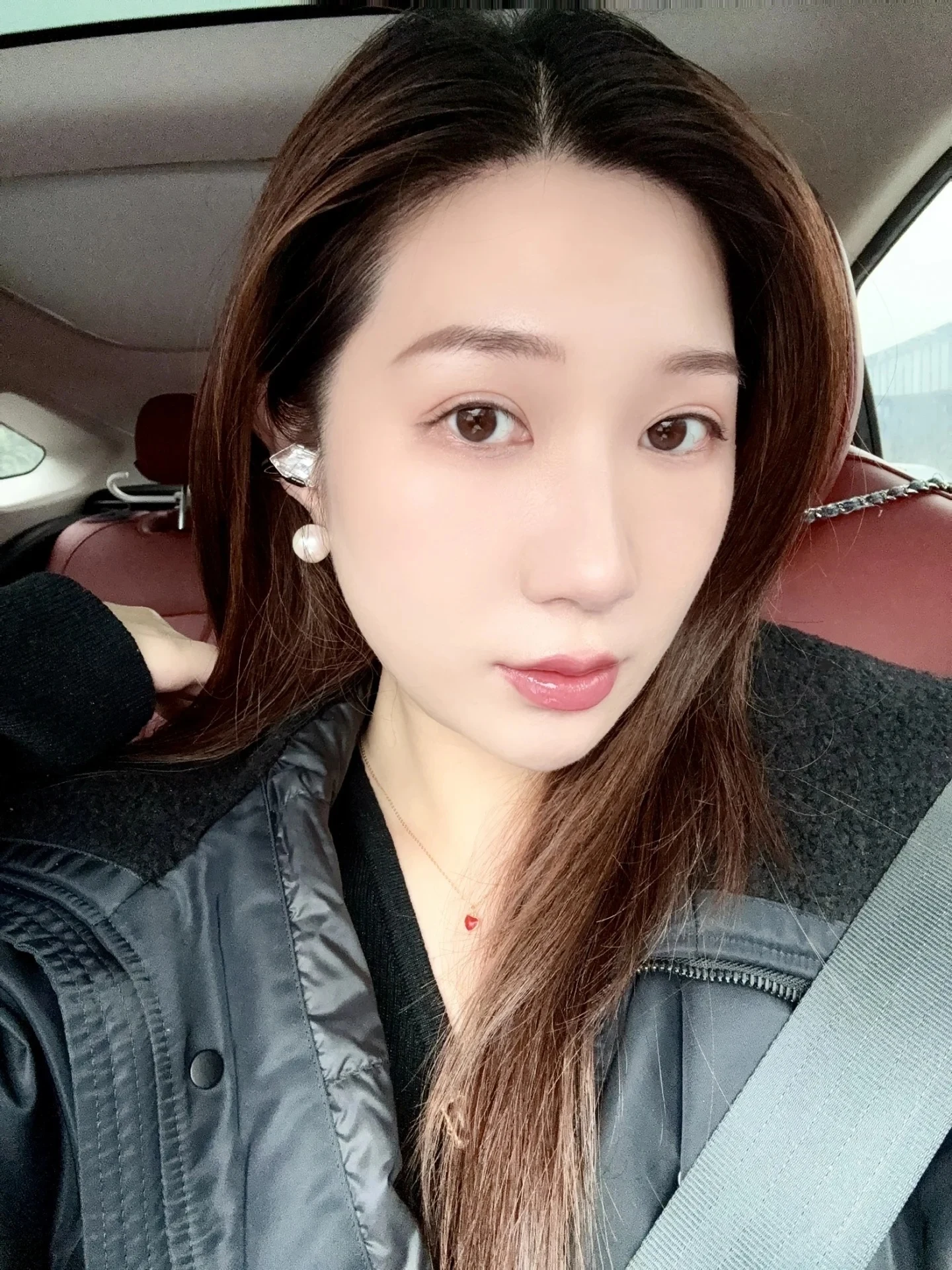 Selfie of a young woman with long brown hair wearing earrings and a jacket inside a car.