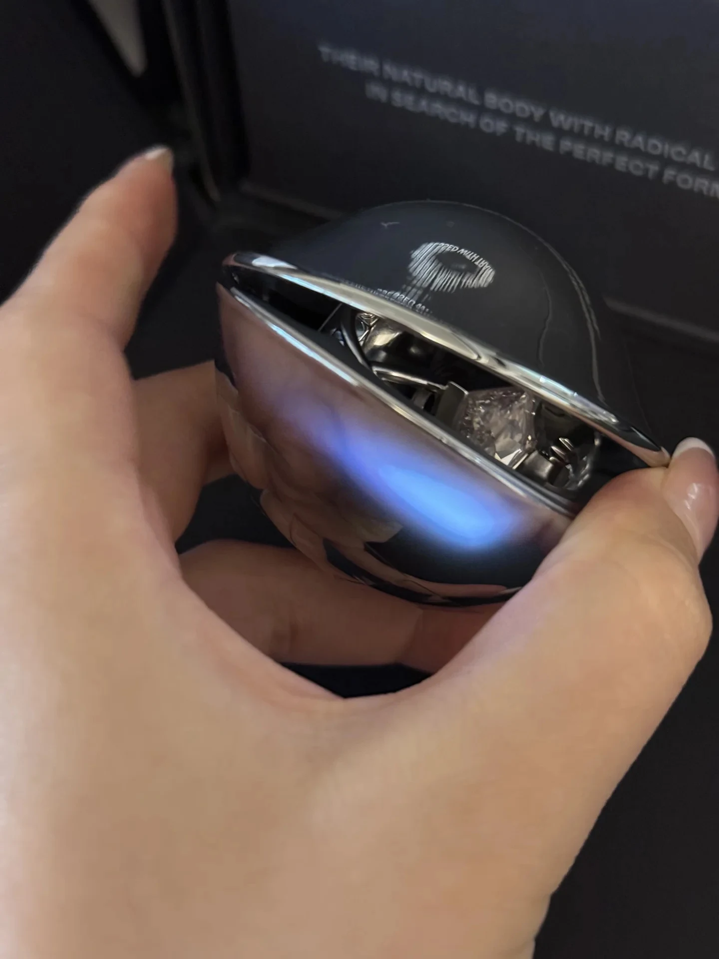 A hand holding a sleek, futuristic spherical capsule or compact case with a metallic chrome finish and a faint blue reflection, slightly open to reveal intricate inner parts.