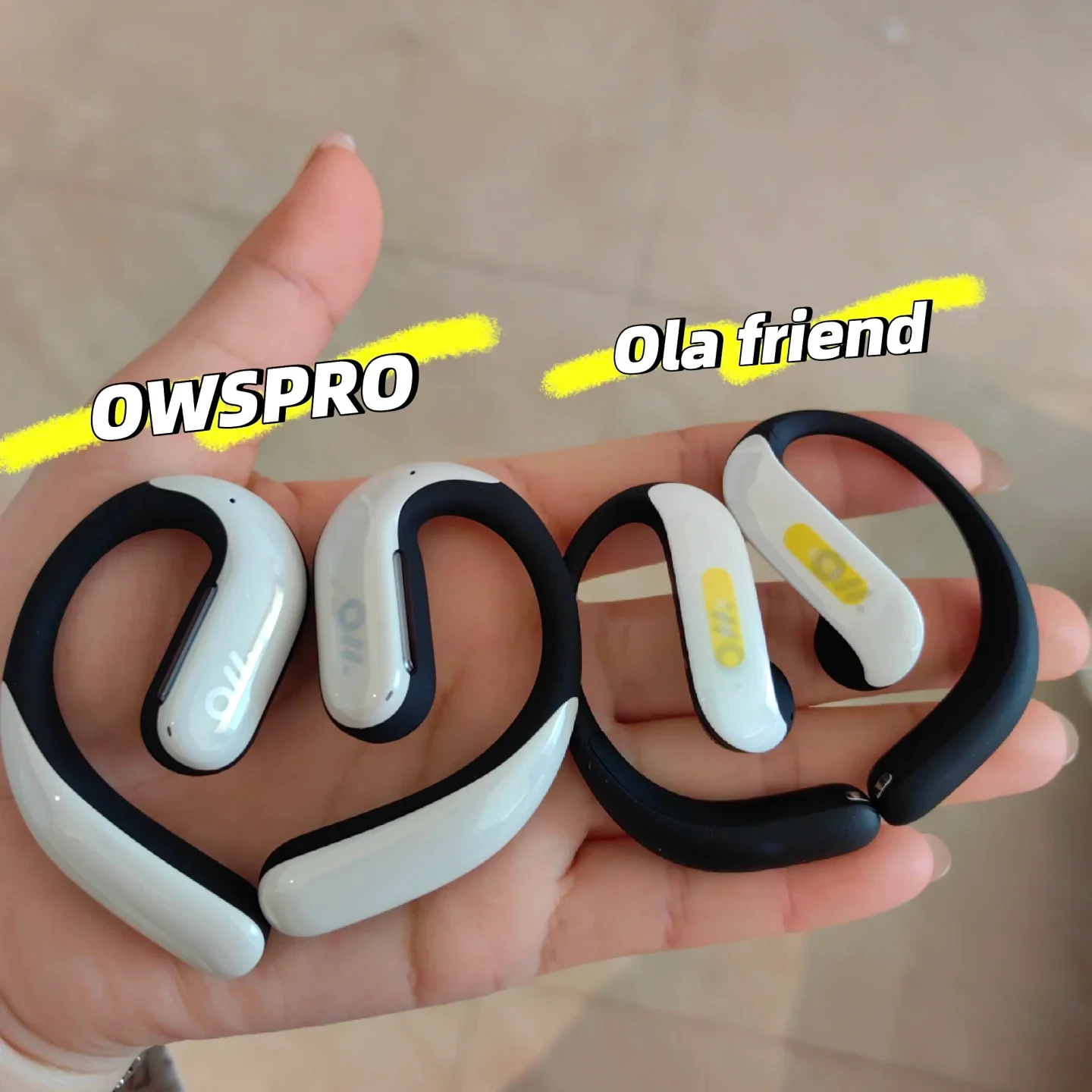 A hand holding two pairs of open-ear clip-on wireless earbuds side-by-side for comparison, with text labels reading "OWSPRO" on the left pair and "Ola friend" on the right pair.