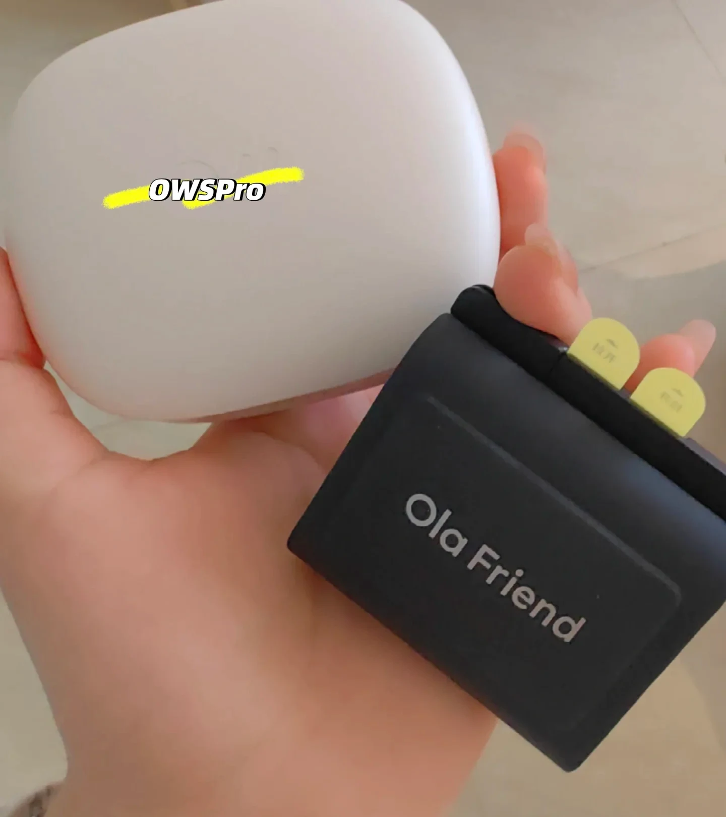 A hand holding two earphone charging cases: a large rounded white case labeled OWS Pro and a smaller black case labeled Ola Friend with yellow pull tabs.