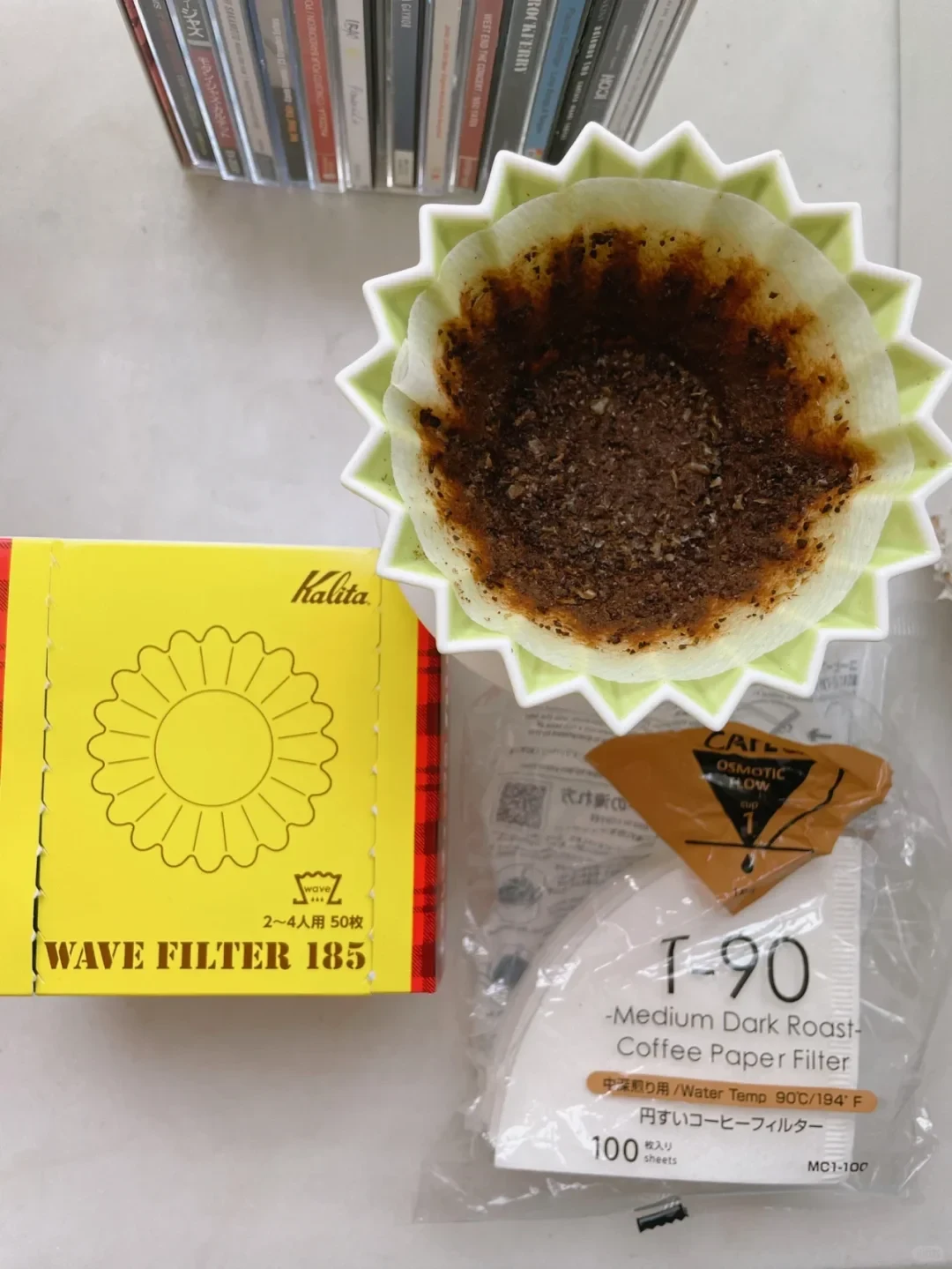 Overhead photo of a coffee dripper holding used grounds beside Kalita Wave Filter 185 and T-90 coffee paper filter packaging.