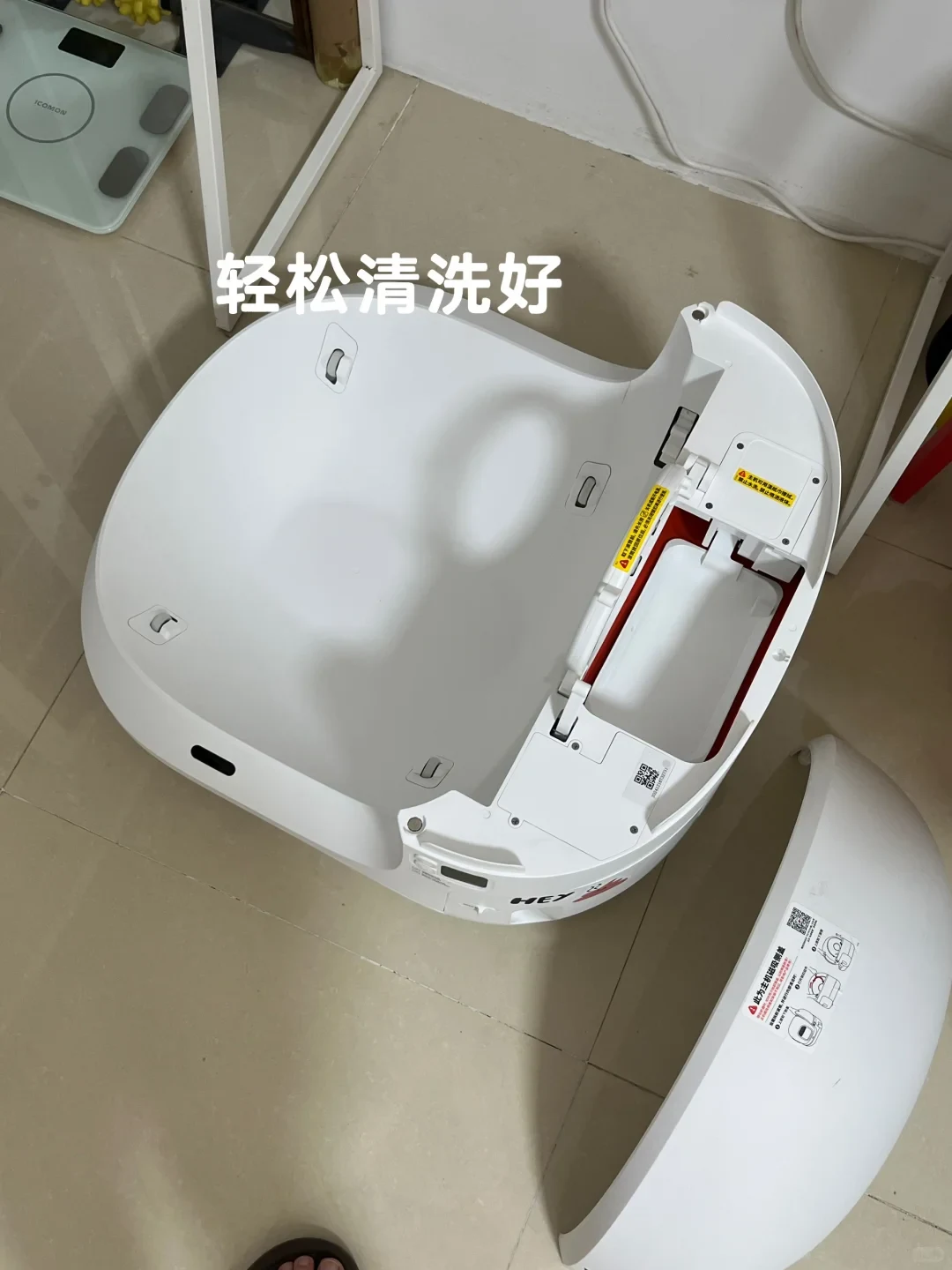 Top-down view of a white automatic pet litter box disassembled on a tiled floor, showing the main basin and its detached outer cover nearby.