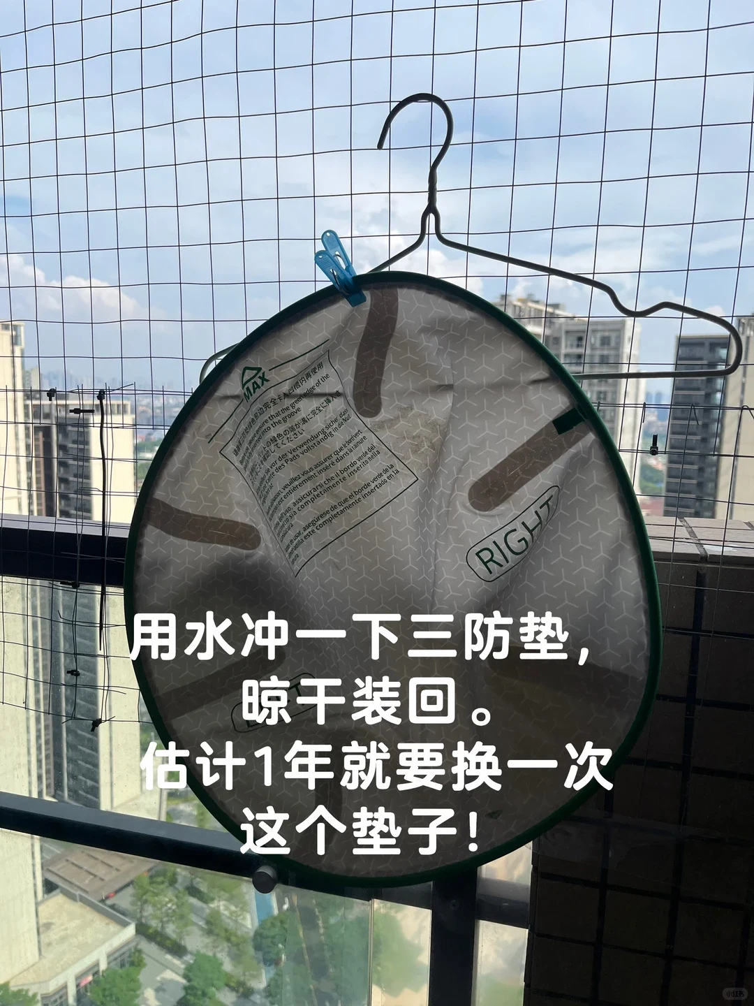 A round liner pad with a green border hanging to dry with a clothespin on a wire hanger outside against a safety net on a high-rise balcony.