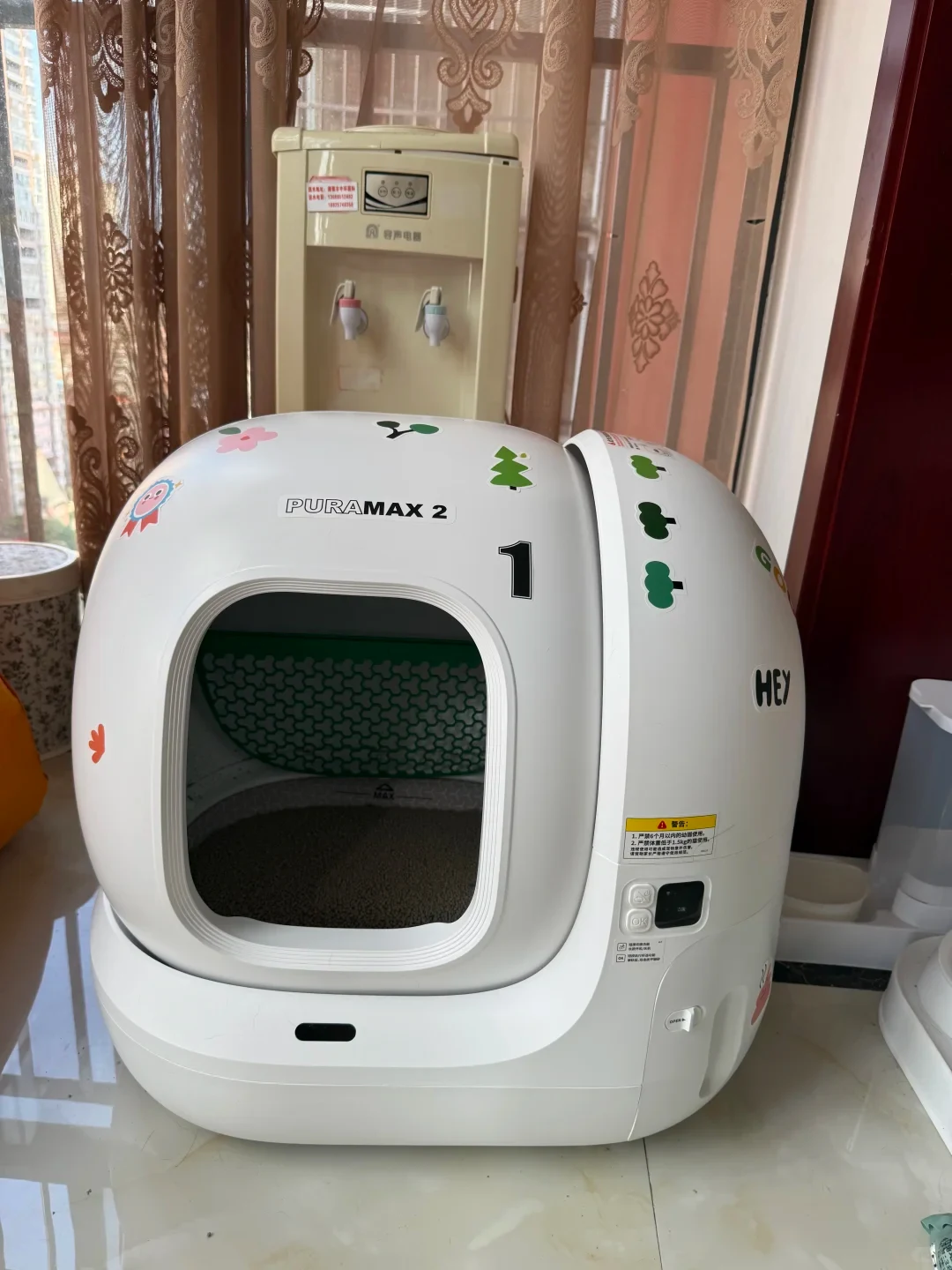 A white PETKIT Pura Max 2 automatic cat litter box decorated with colorful stickers, placed on a tiled floor indoors with cat litter visible inside the front drum opening and control buttons on the side panel.