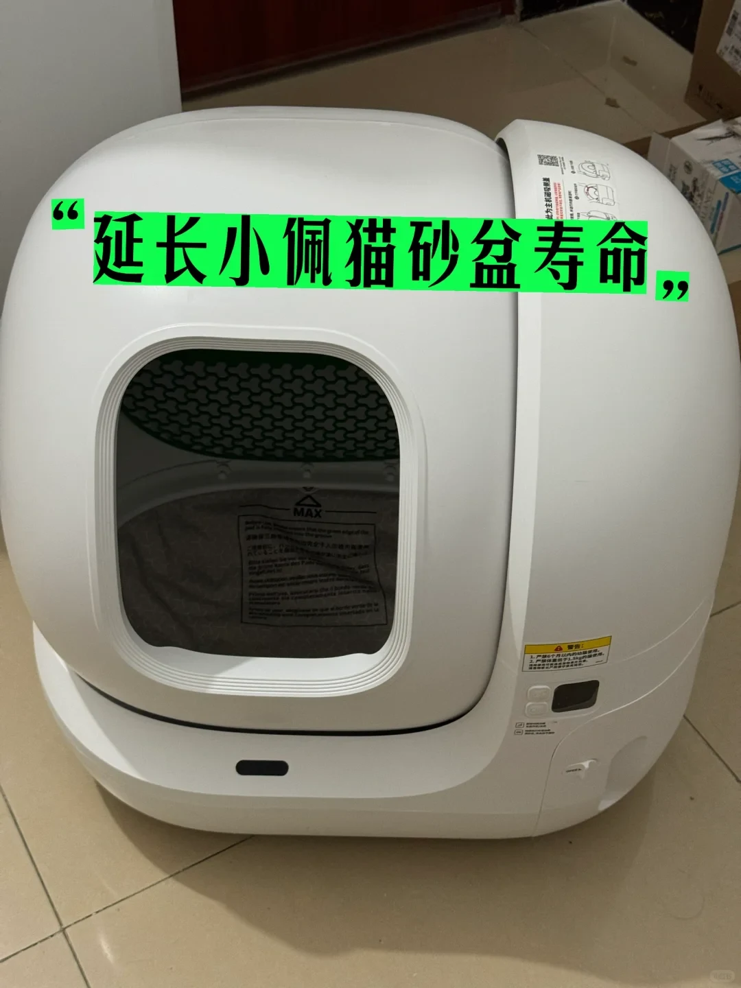 A white PETKIT automatic cat litter box on a tiled floor with a front opening into the drum, control panel on the right, and overlaid Chinese text across the upper front.