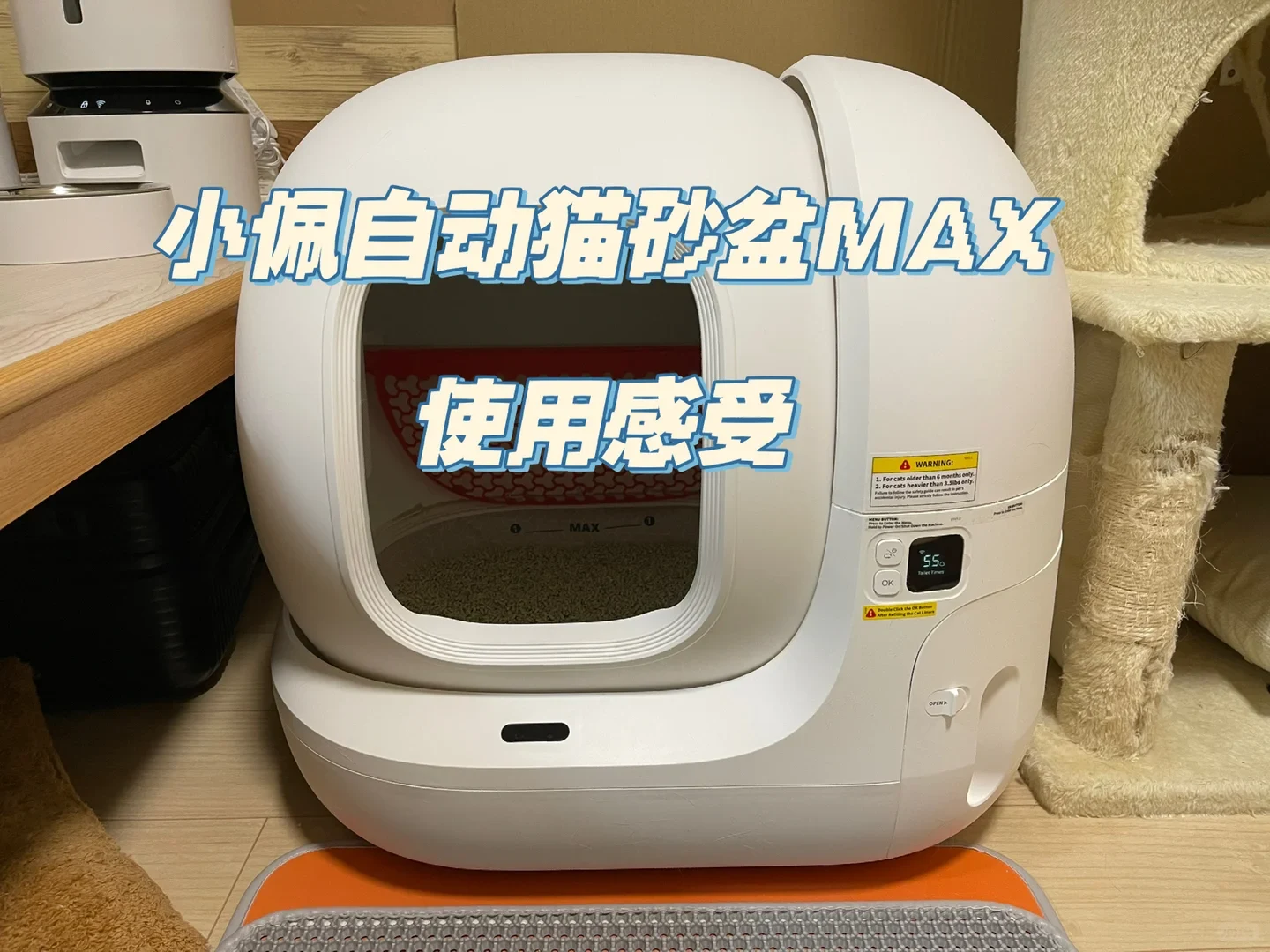 A white Petkit automatic self-cleaning cat litter box with litter inside, featuring an entryway, small digital control display, and warning labels, set on the floor between a pet feeder and a cat tree.