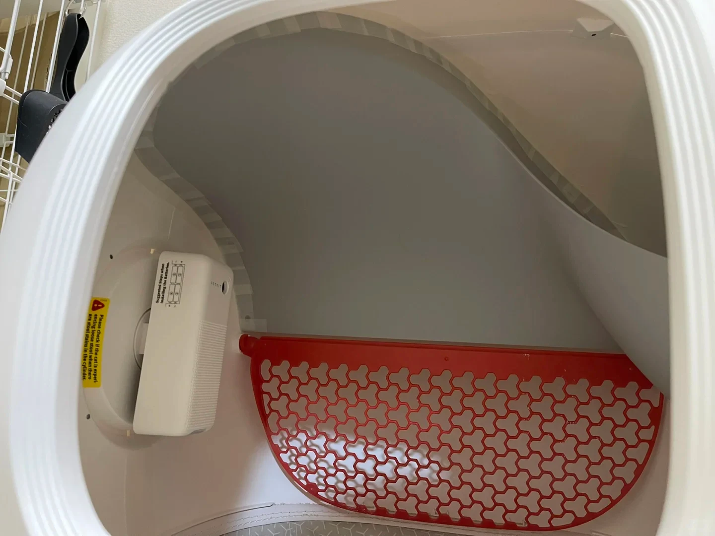 Interior view of an automatic cat litter box chamber showing a white deodorizer unit mounted on the left inner wall, a yellow warning label, and a curved red plastic sifting grate set against the grey curved interior.