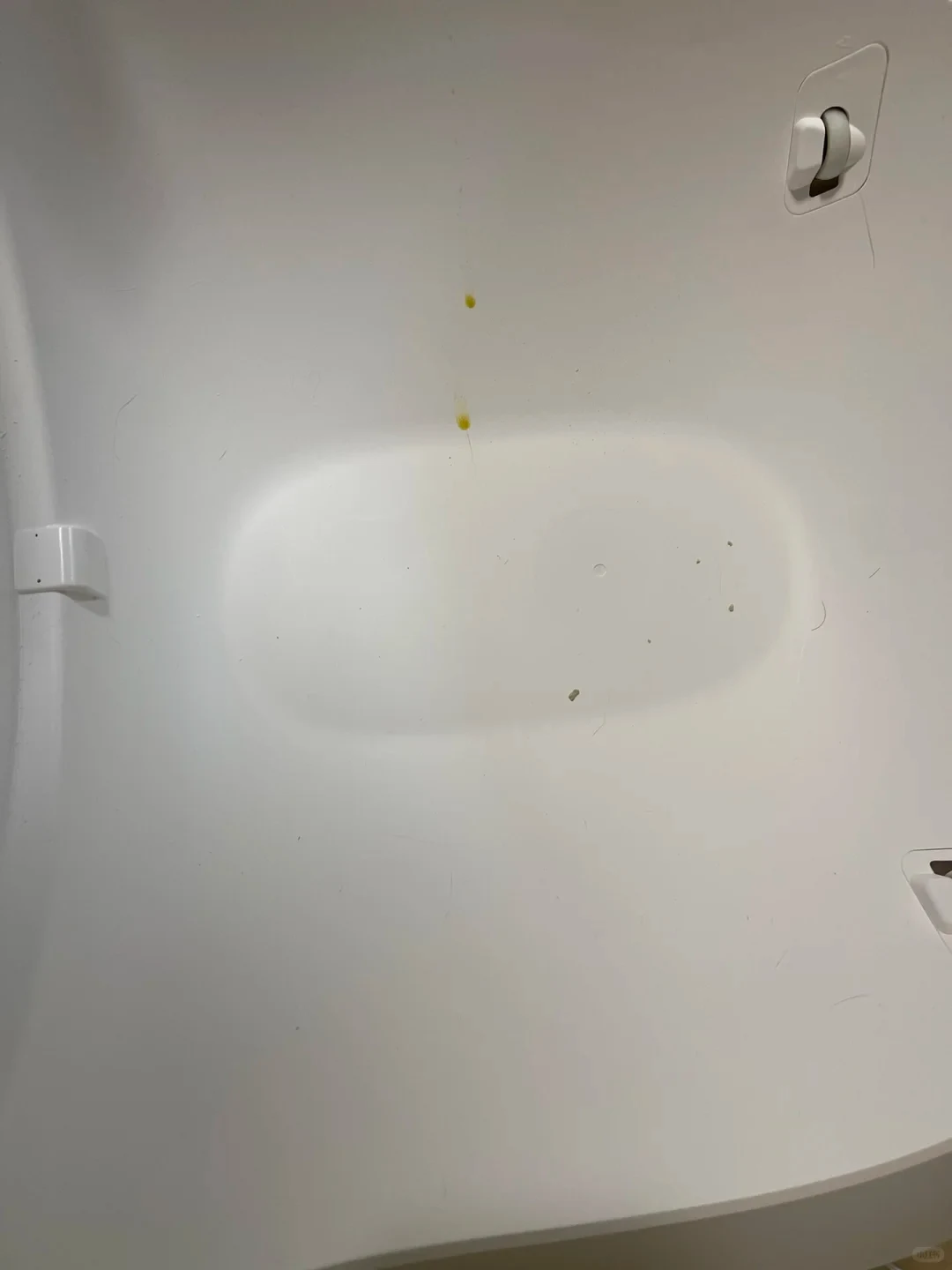Interior view of a white plastic basin or appliance compartment, featuring yellowish liquid droplets dripping down the curved wall into the central recessed area, with stray hairs and small specks of dirt on the surface.