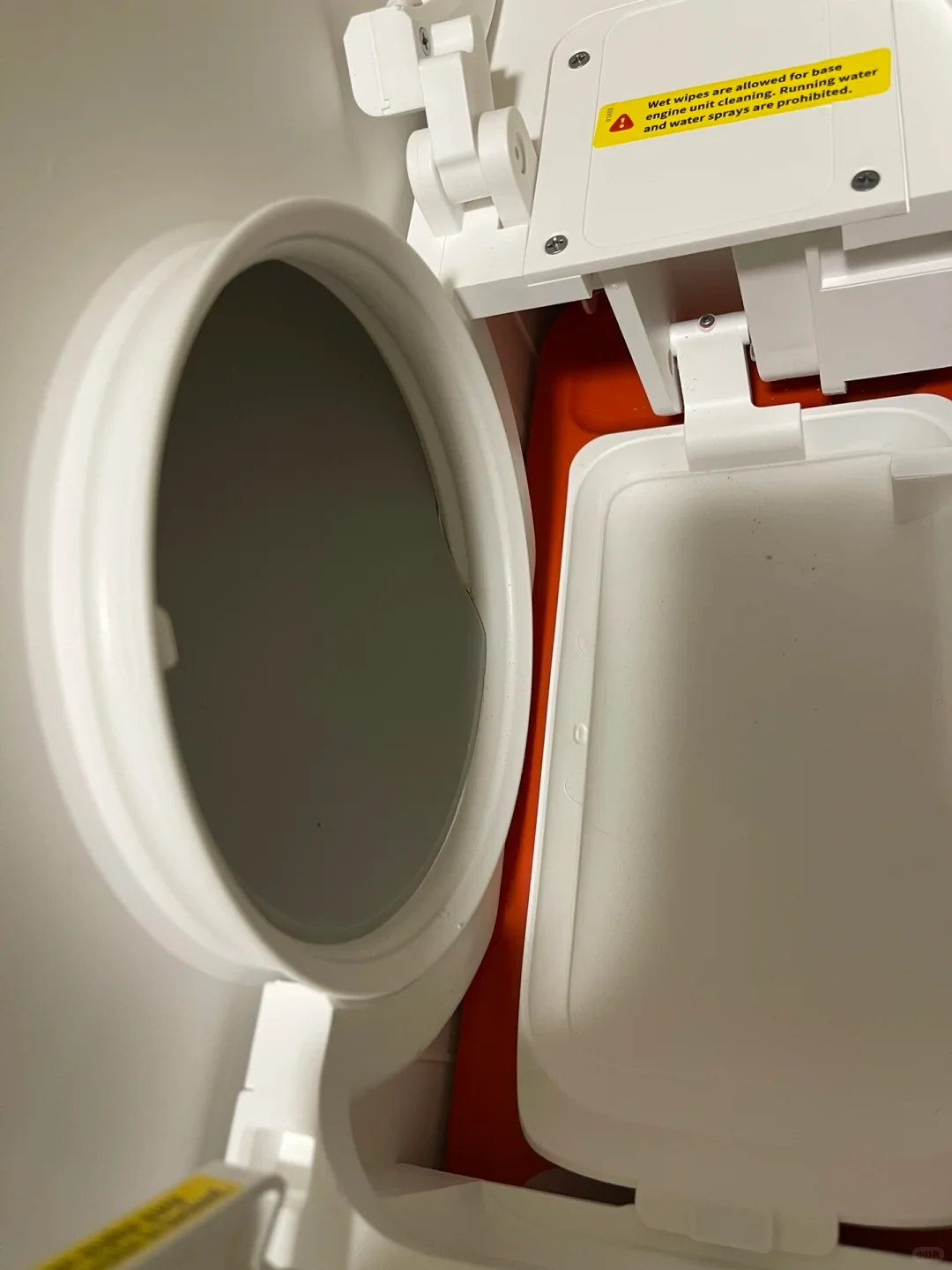 Interior mechanical compartment of a white plastic appliance with a circular aperture, hinged access covers, and a yellow warning sticker reading 'Wet wipes are allowed for base engine unit cleaning. Running water and water sprays are prohibited.'