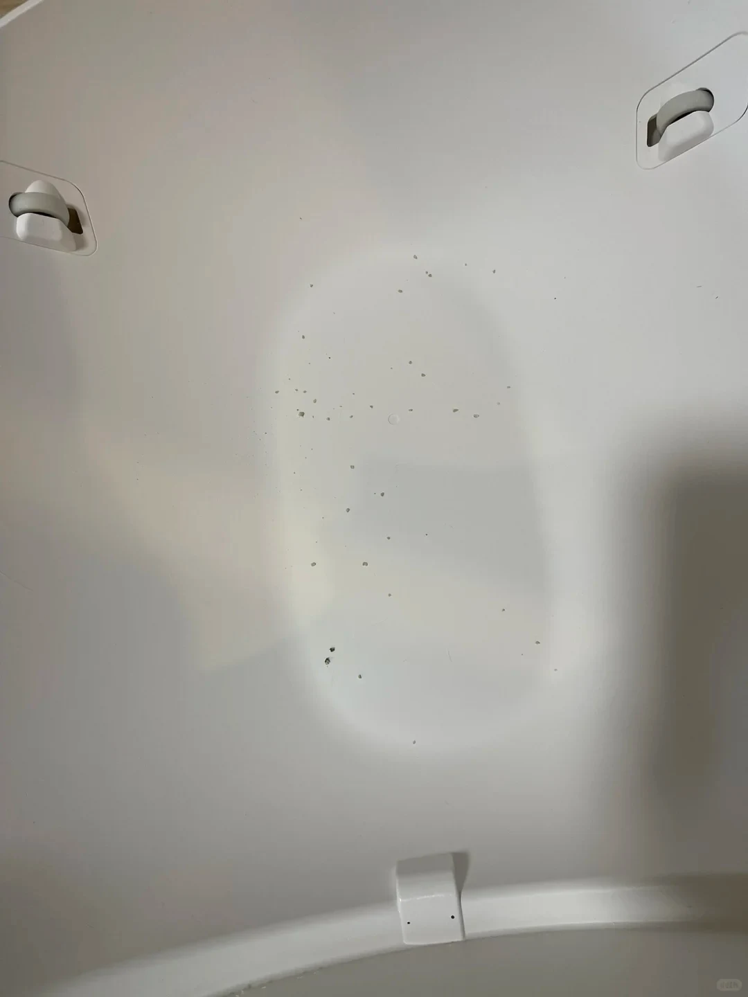 A close-up view of the interior surface of a smooth, white curved plastic appliance, likely a cat litter box drum, with scattered small grey litter particles in the center and mechanical clips visible along the edges.