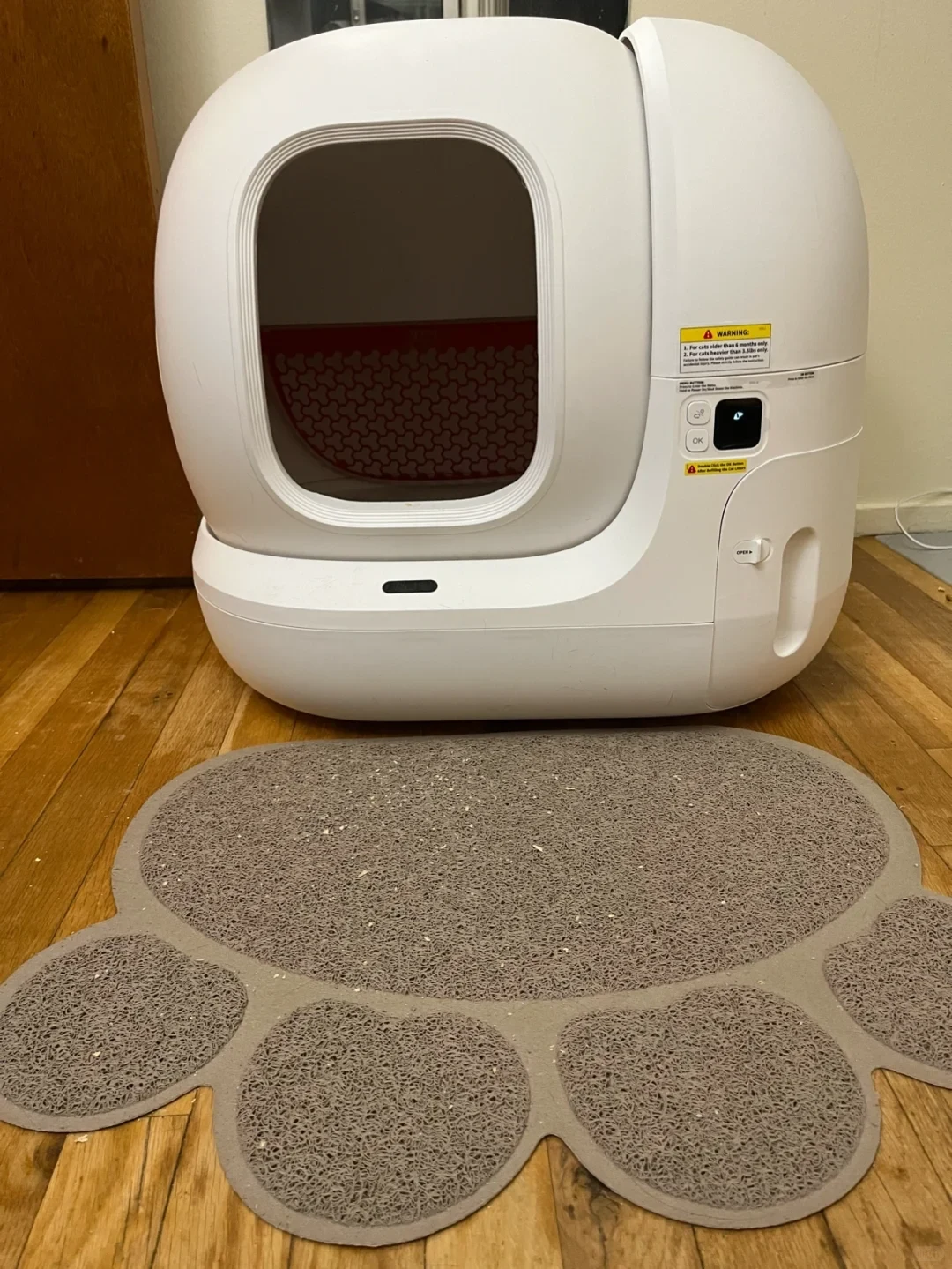 A white automatic self-cleaning cat litter box on a wooden floor, with a paw-shaped grey textured litter trapping mat placed in front of its entrance.