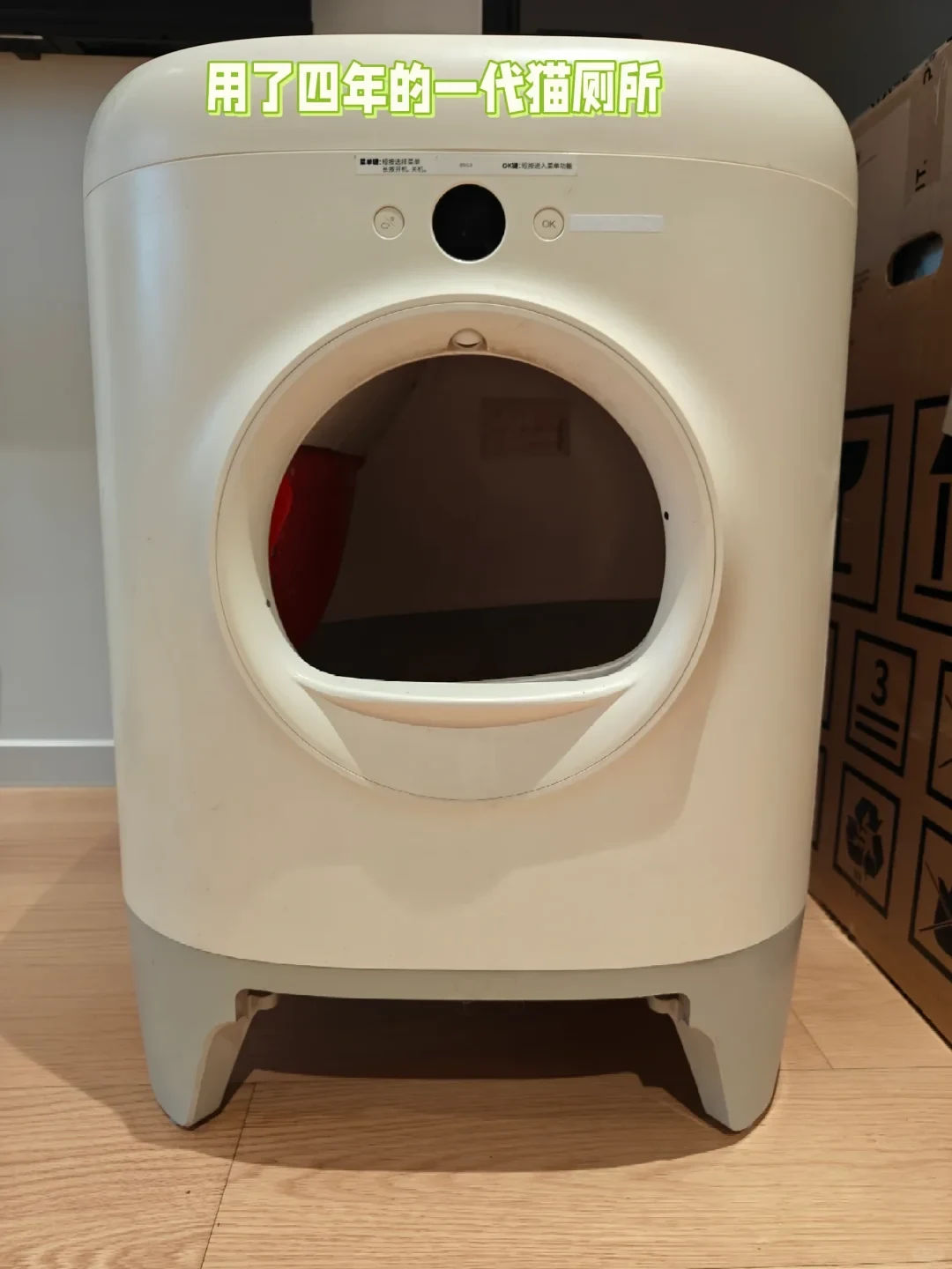 Front view of an off-white smart automatic cat litter box with a large circular entryway, standing on a light wood floor next to a cardboard box.