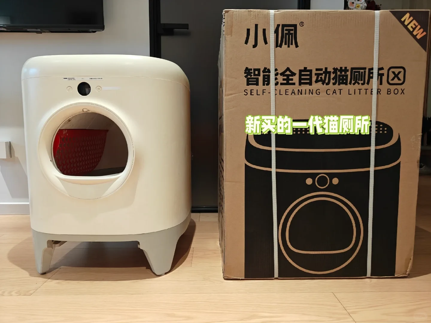 An unboxed white automated self-cleaning cat litter box stands next to its large cardboard packaging box on a light wood floor indoors.