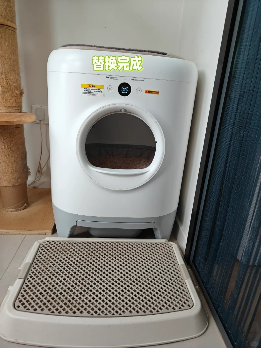 A white automatic self-cleaning cat litter box positioned on the floor next to a sisal cat scratching post. The machine features a circular entrance showing cat litter inside, a circular digital screen reading 100%, and a beige textured litter-trapping mat placed directly in front on the tiled floor.