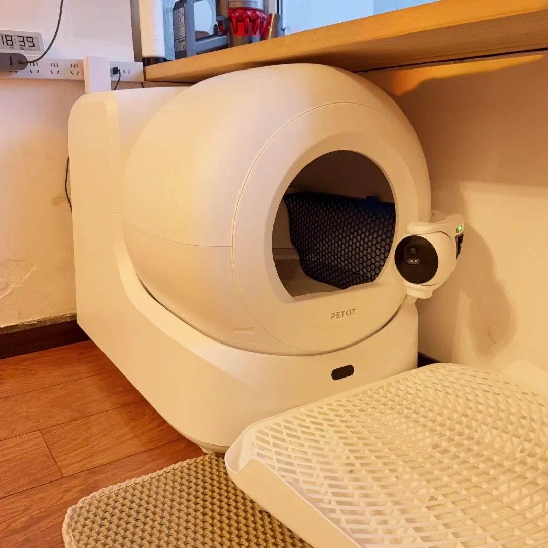 A white PETKIT Ultra automatic cat litter box under a wooden desk with its camera, a litter-control board and a separate mat in front.