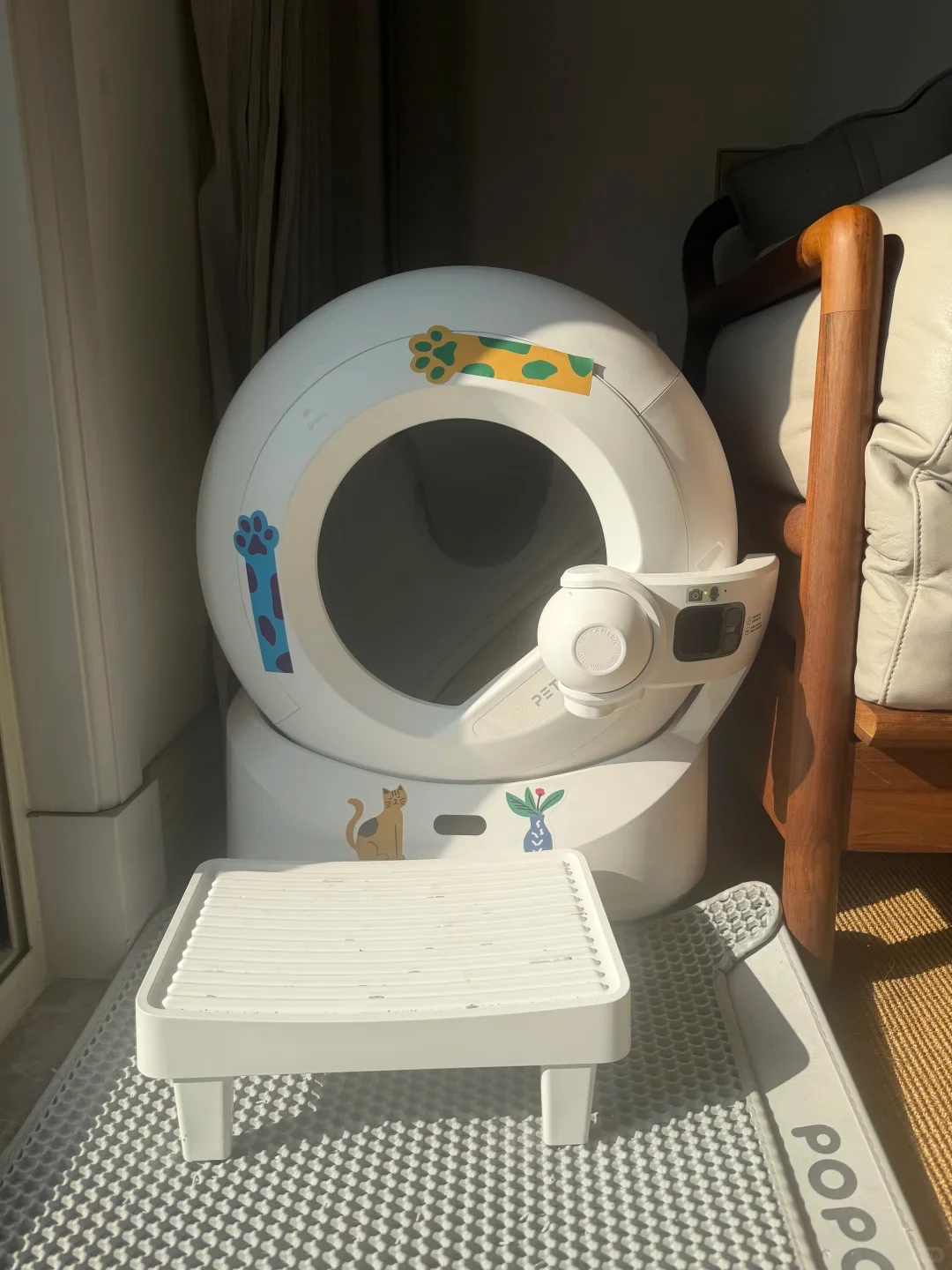 A white Petkit Ultra automatic litter box with a front-mounted camera module, decorated with stickers, positioned behind a white step stool on a textured mat beside a chair.
