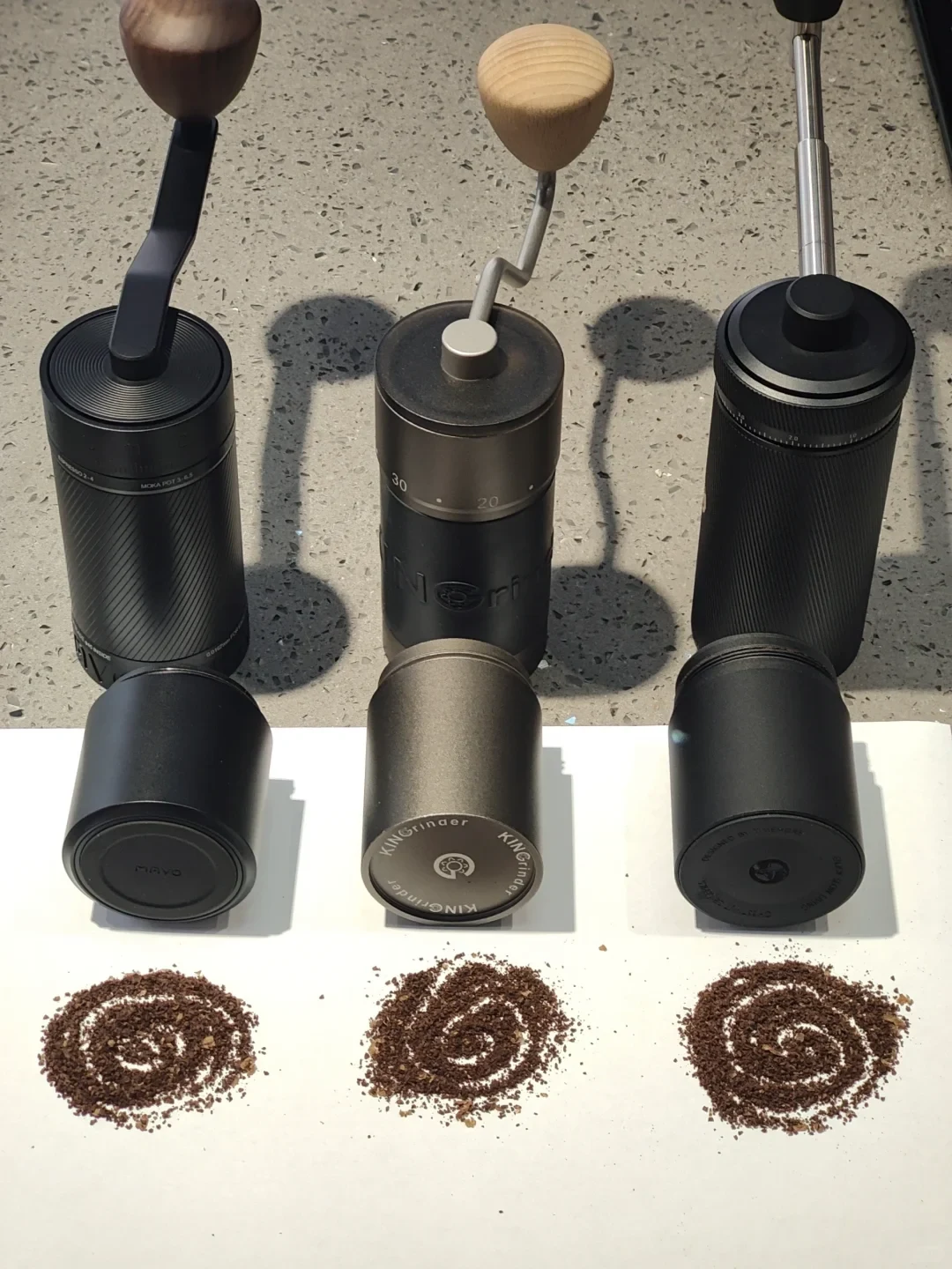 Three manual coffee grinders stand side by side on a countertop, each with a different handle design. Their detached catch cups sit in front, above three piles of ground coffee arranged in spiral patterns.