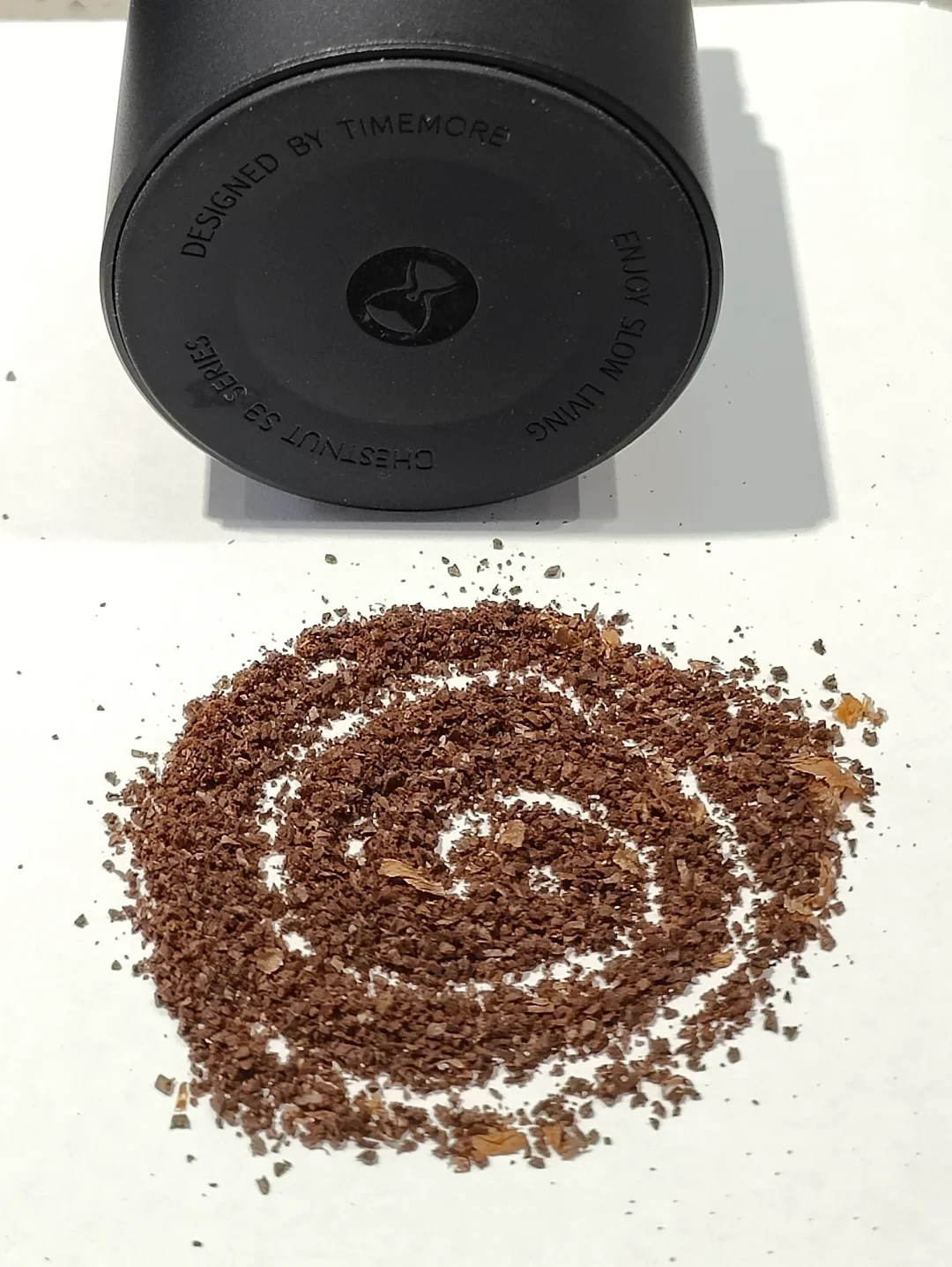 The base of a detached TIMEMORE Chestnut S3 catch cup, marked “CHESTNUT S3 SERIES,” above coffee grounds arranged in a spiral.
