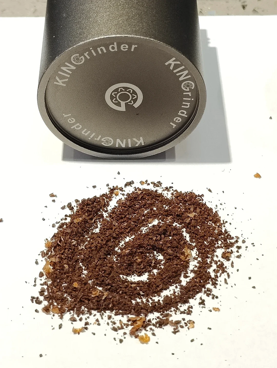 A detached KINGrinder catch cup with its branded base visible above coffee grounds arranged in a spiral on a white surface.