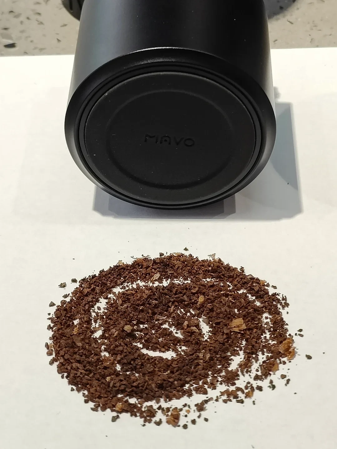 The MAVO-branded base of a detached catch cup above coffee grounds arranged in a spiral on a white surface.