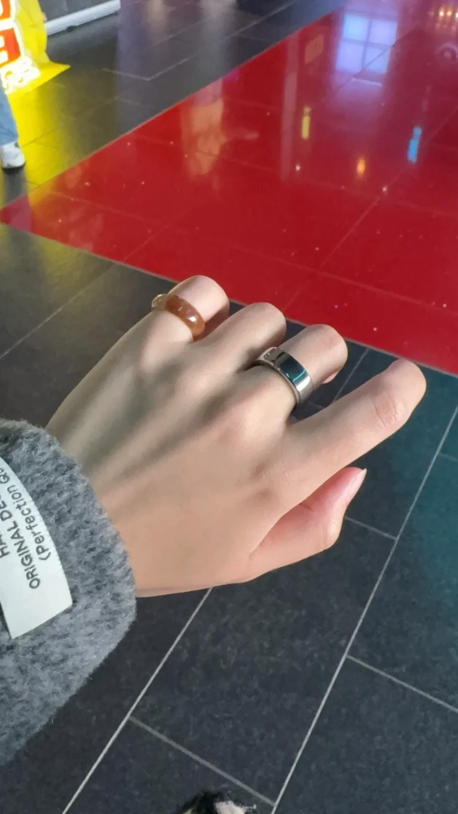 A close-up view of a hand wearing an amber-colored resin ring on the index finger and a wide silver band ring on the middle finger, set against a background of red and dark grey floor tiles.