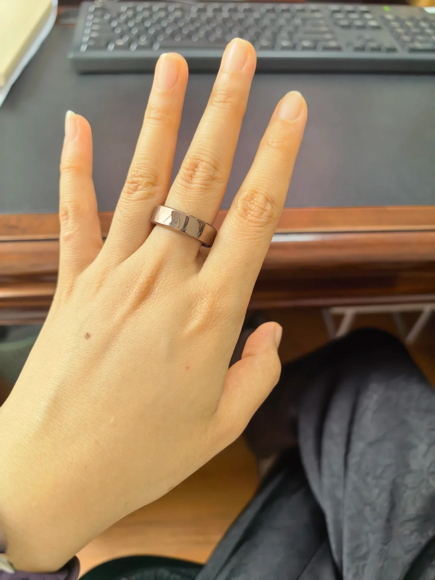 A hand wearing a wide, metallic smart ring on the middle finger, held over an office desk with a keyboard visible in the blurred background.