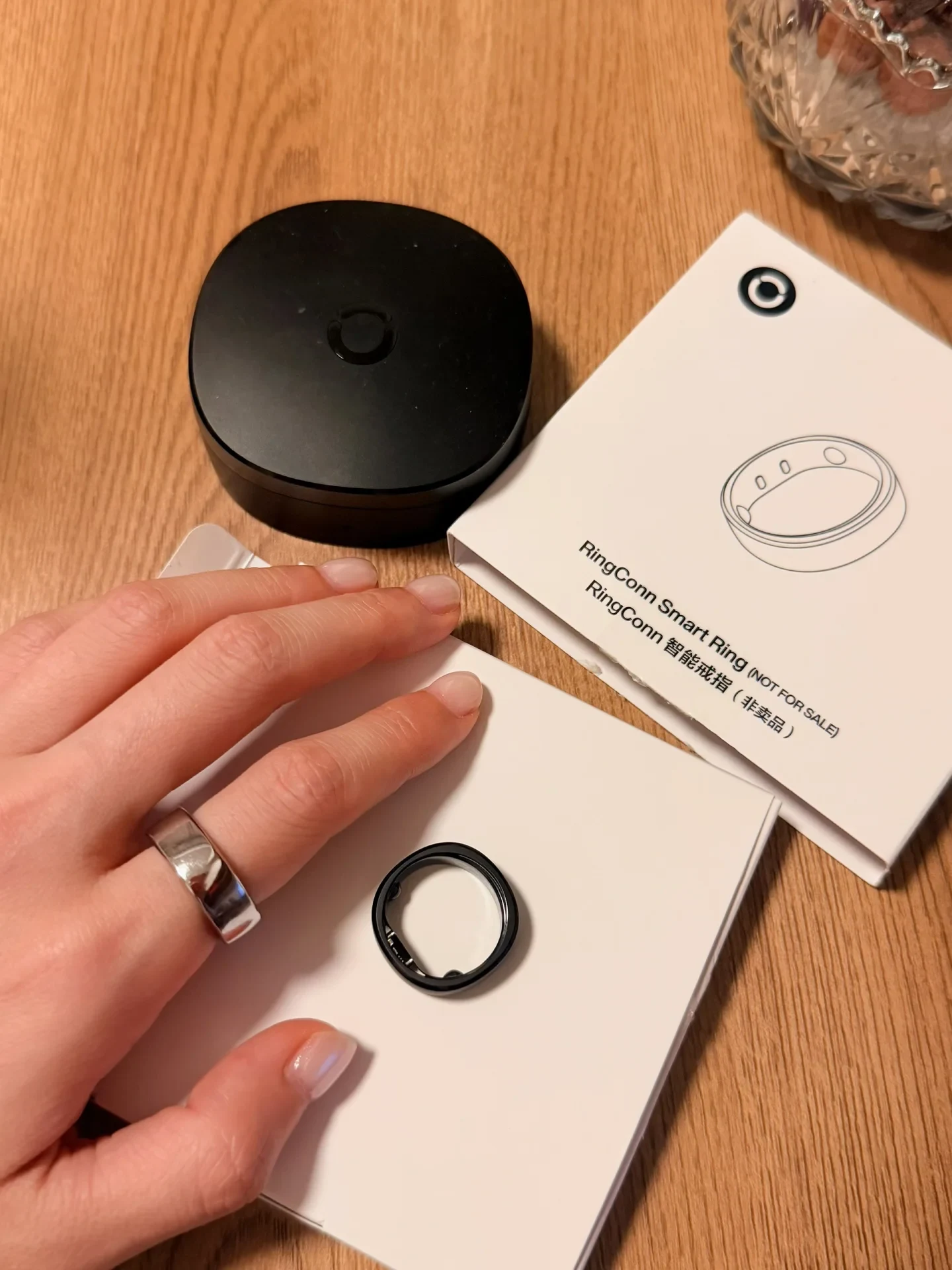 A hand wearing a silver smart ring rests beside an open white box containing a black smart ring, alongside a black charging case and outer packaging.