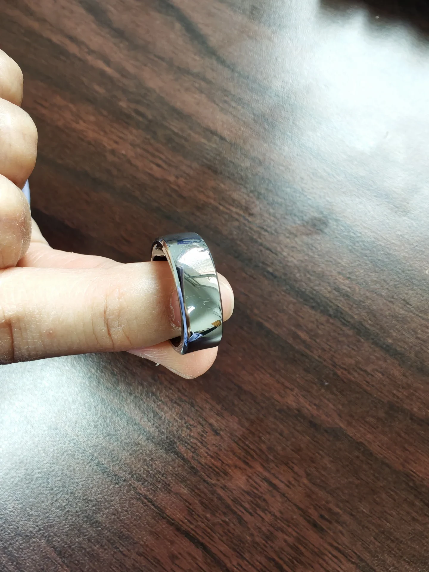A close-up view of a glossy dark metallic smart ring worn on a person's finger over a dark wood table.