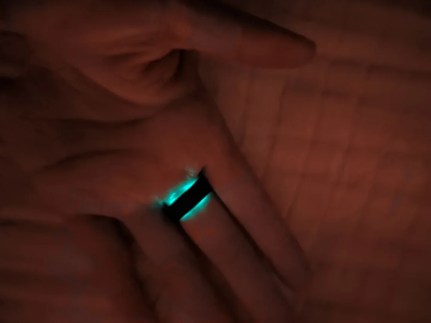 A close-up of a hand wearing a black ring that glows with bright turquoise light along its inner edges in dim red lighting.