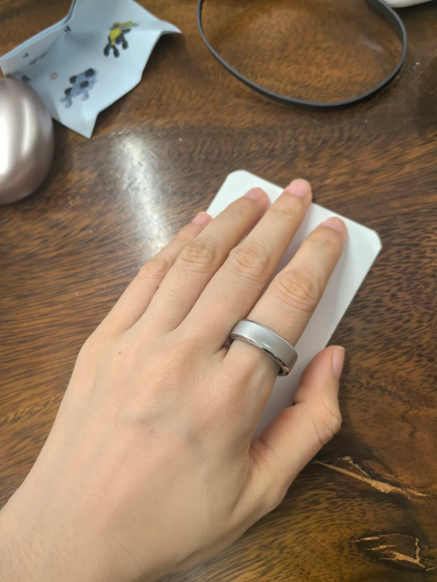 A hand wearing a silver-toned smart ring on its ring finger rests atop a white device on a wooden desk.