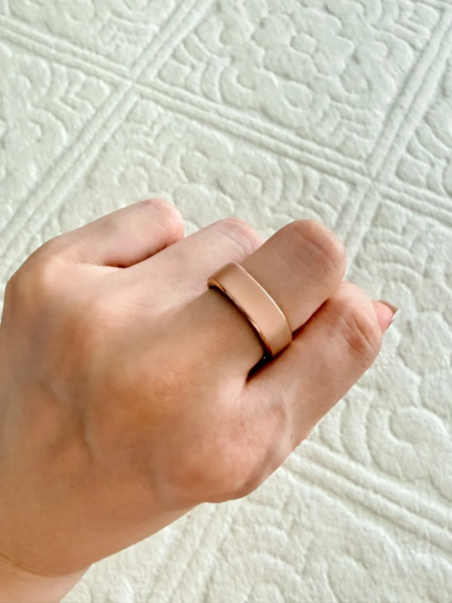 A hand wearing a wide, smooth rose-gold band ring on the index finger, resting over a textured off-white quilted surface.