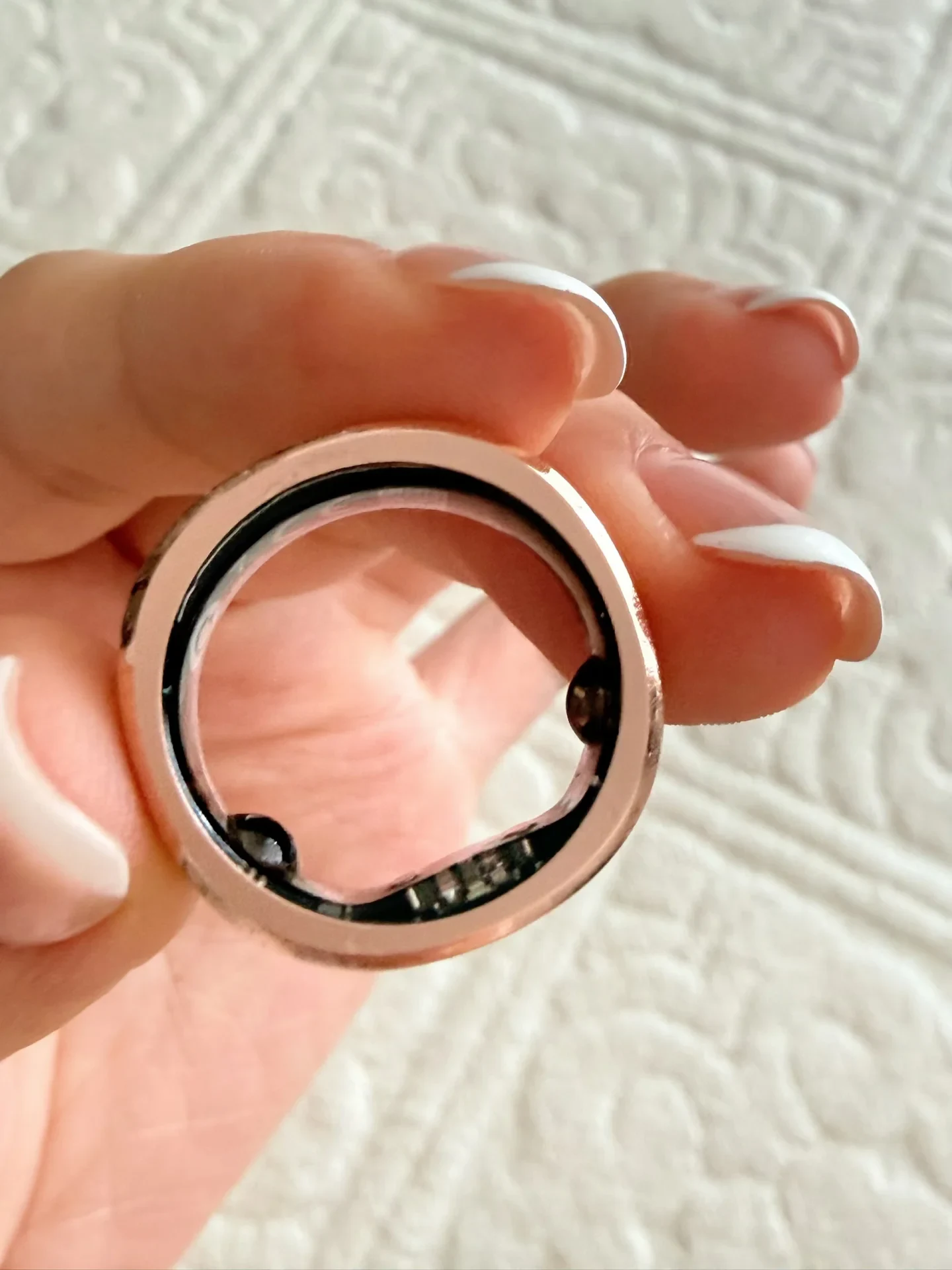 A hand with white-tipped manicured nails holds up a rose gold smart ring, showing the internal sensors on the inner band.