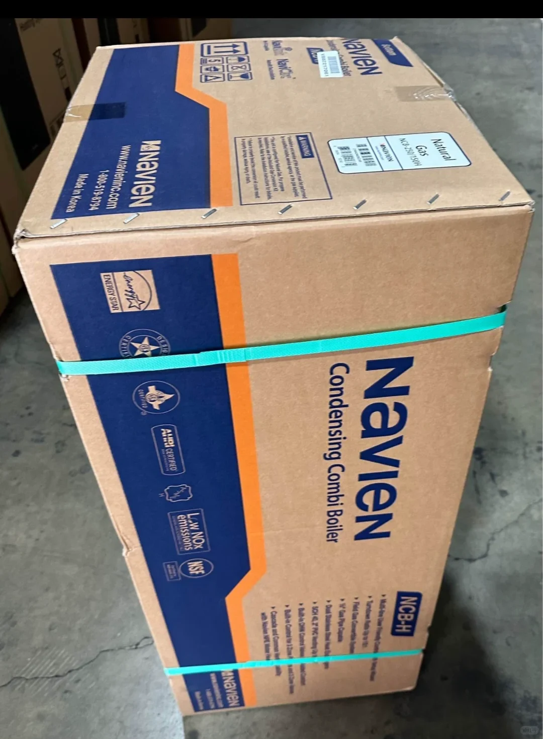 A Navien condensing combi-boiler carton secured with turquoise straps. The label reads NCB-250/150H and Natural Gas.