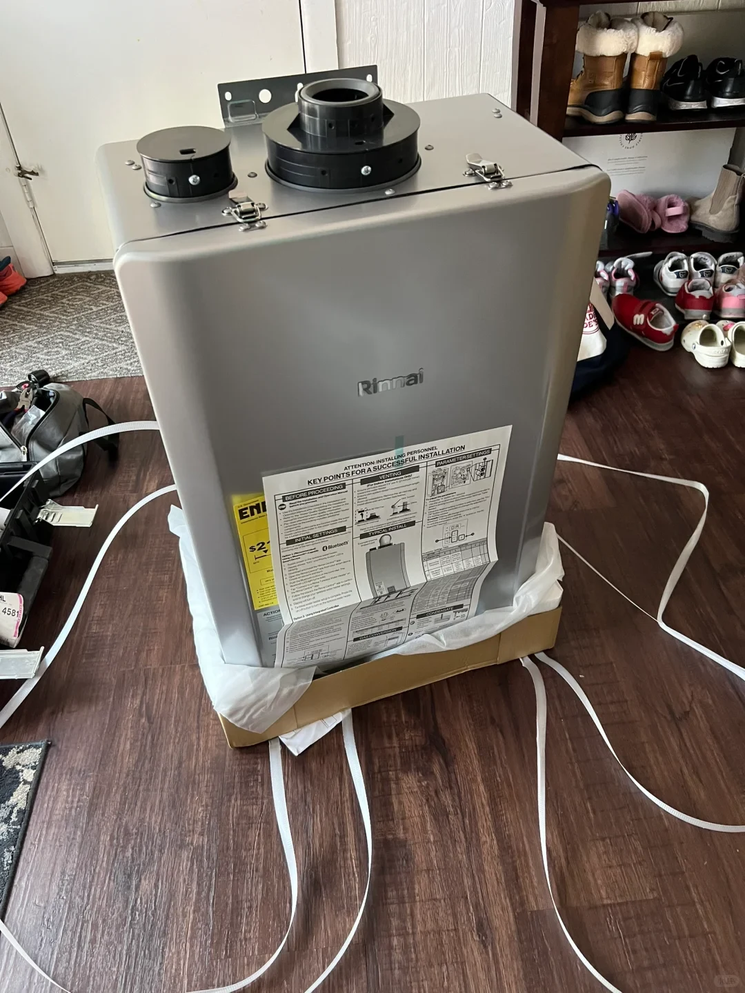 A gray Rinnai appliance, apparently a tankless water heater, sits partly unpacked in a cardboard box with plastic wrapping. Two black circular vent fittings are mounted on top, and printed installation instructions are attached to the front. Shoes and other household items are visible in the background.